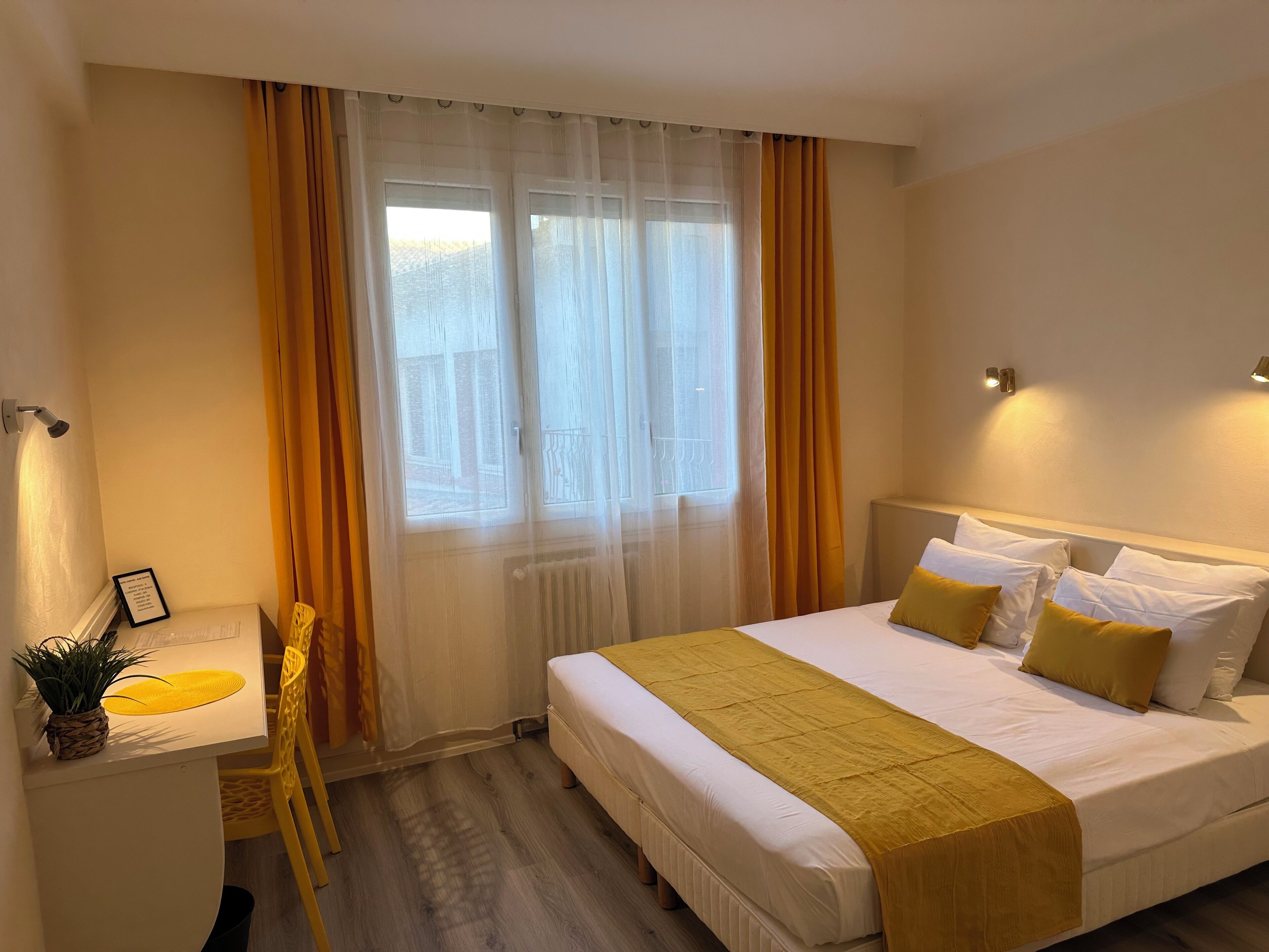 A hotel room with a bed, bedside tables, a chair, a potted plant, and a window with curtains.