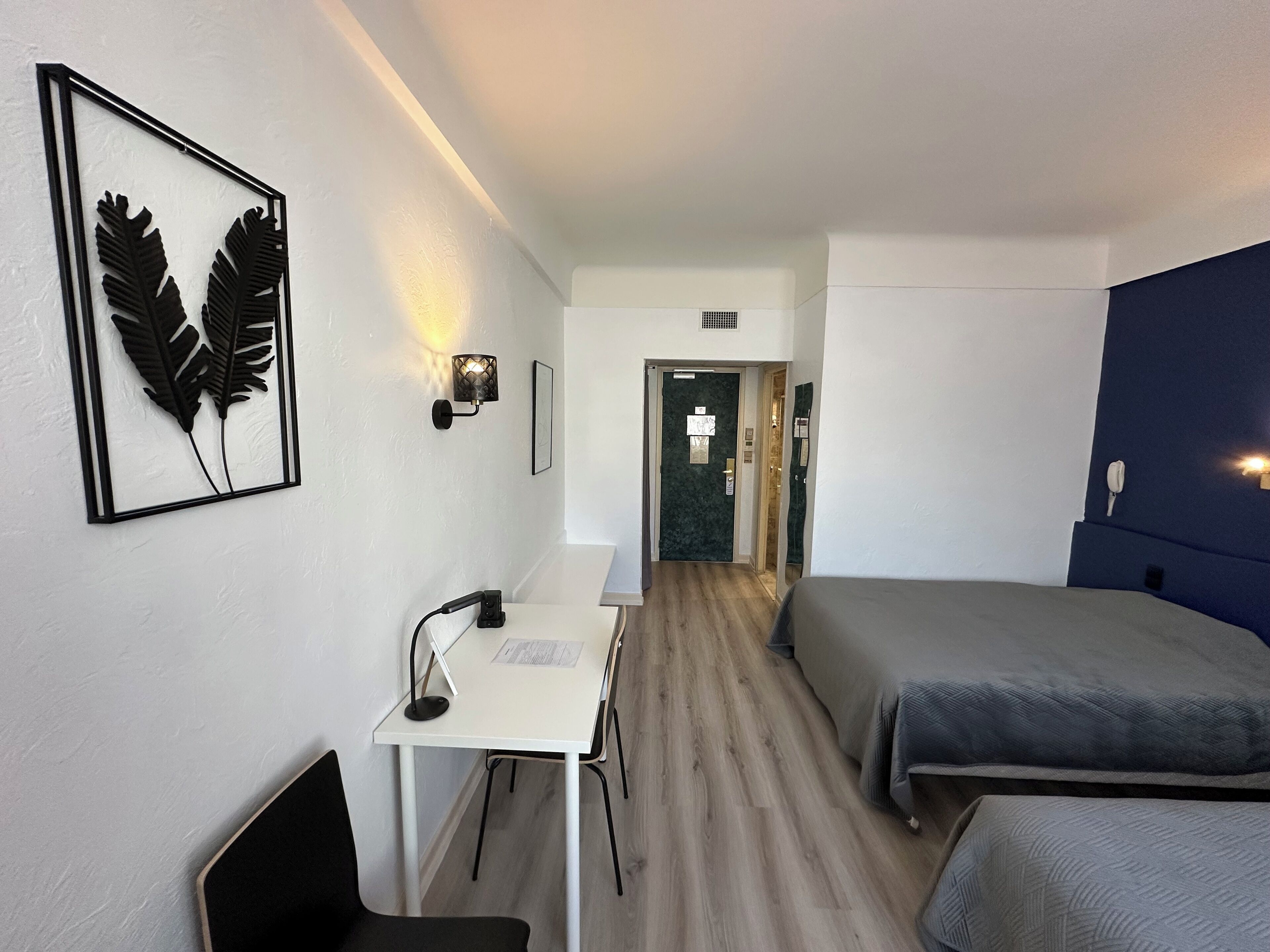 Hotelkamer met een bureau, twee bedden, een lijst met een afbeelding van een blad, een wandlamp en een deur naar een badkamer.