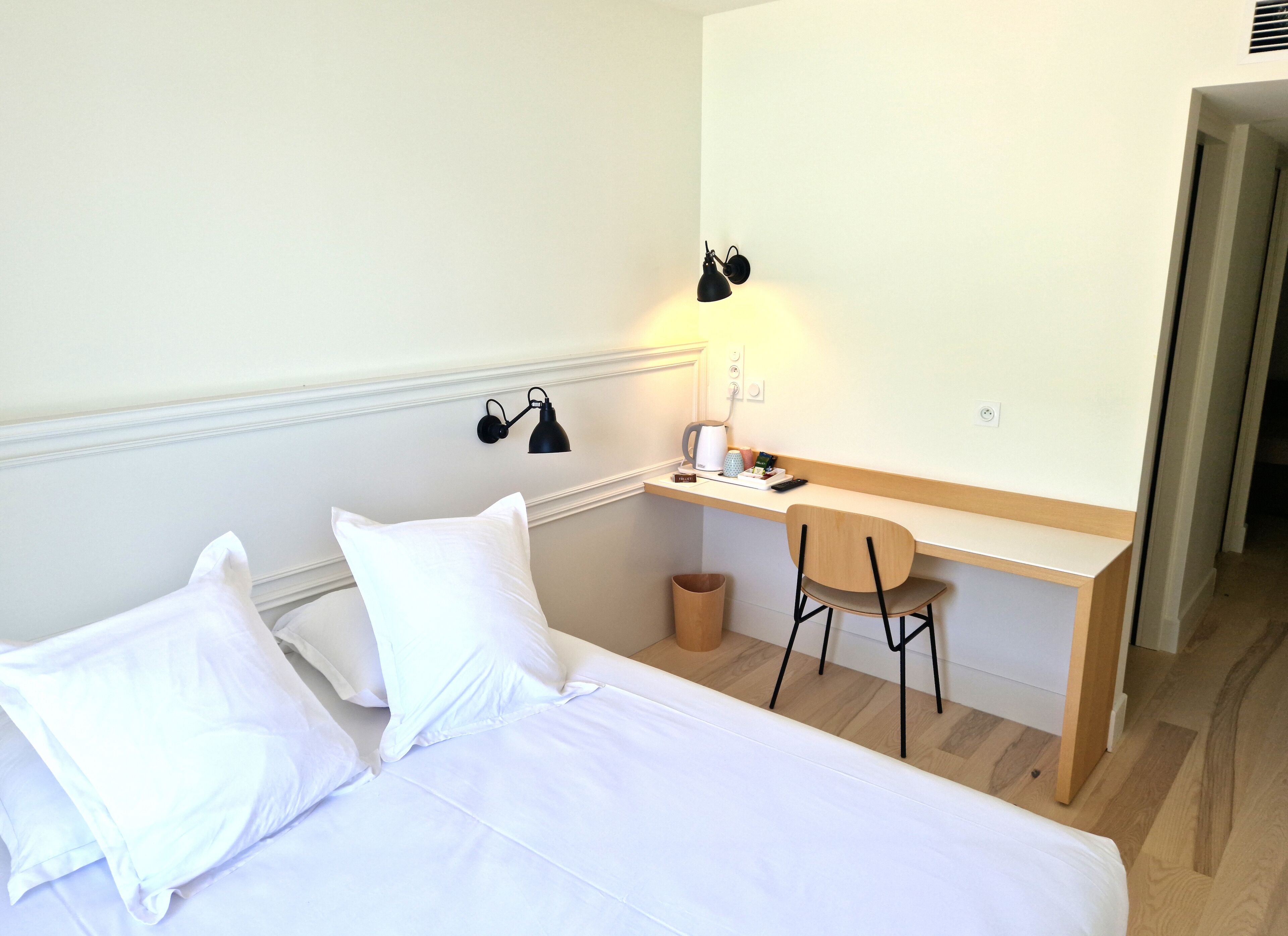 Ett hotellrum med en säng, ett skrivbord, en stol, en lampa och en papperskorg.