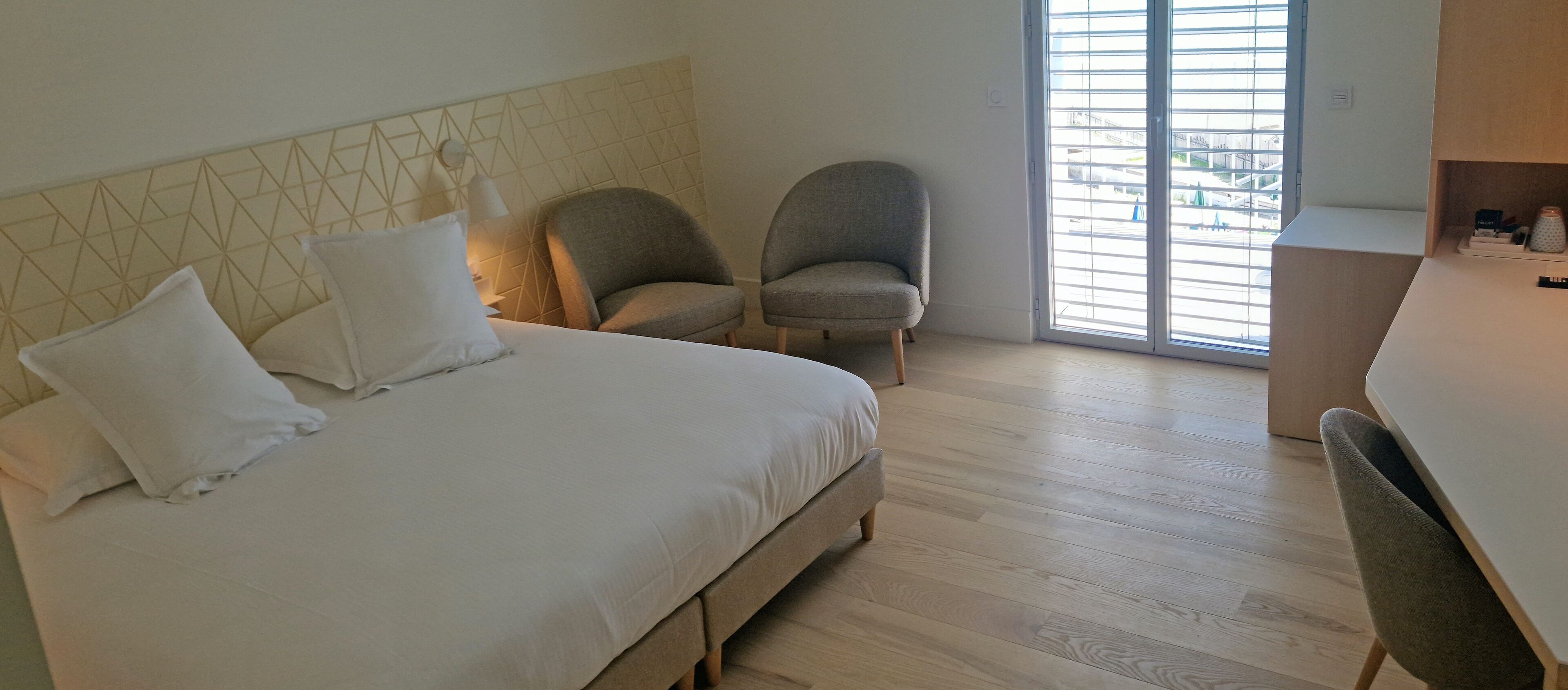 A bedroom with a bed, two armchairs, a desk, and a window with blinds.