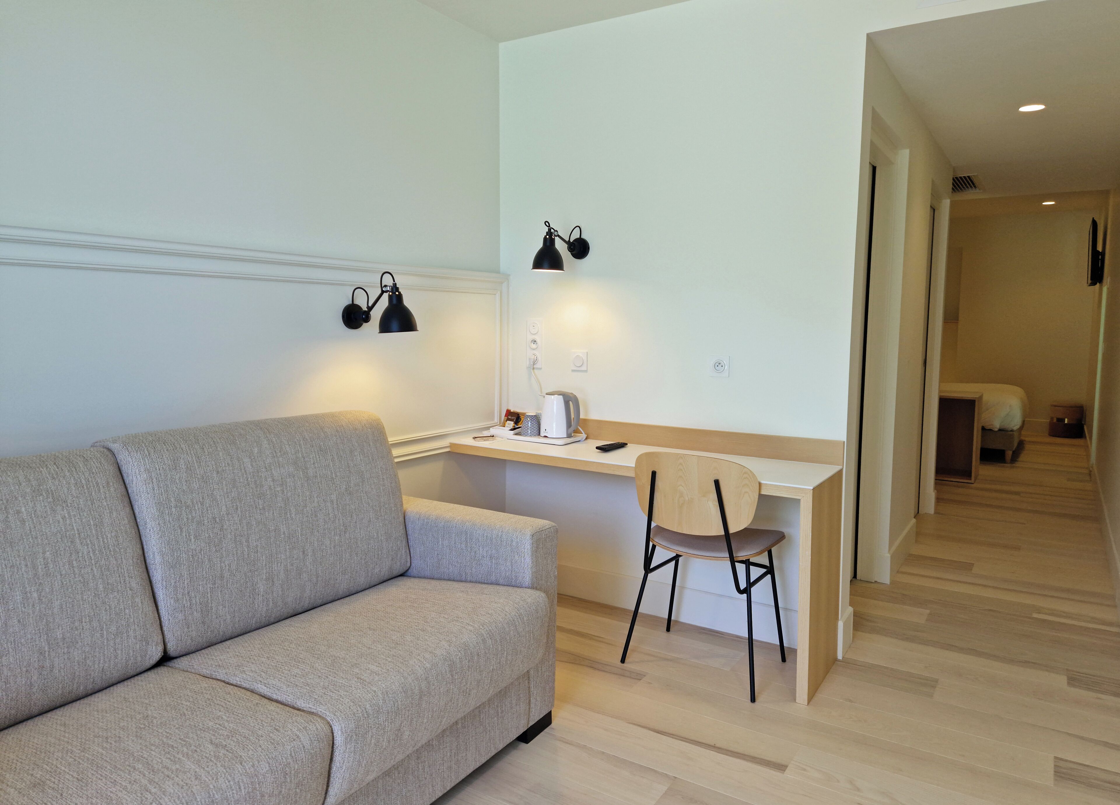 Una habitación moderna con sofá, escritorio y silla, lámpara y cama al fondo.