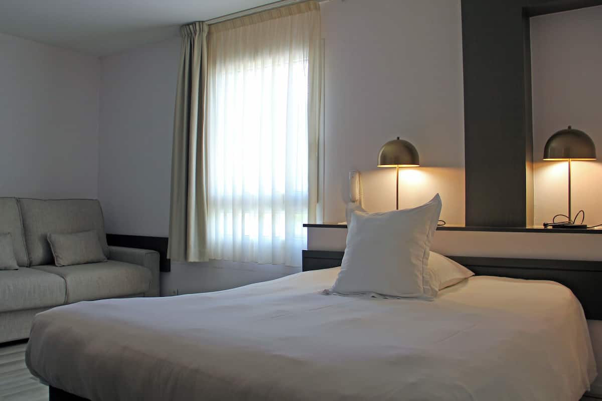 A hotel room with a large bed, a sofa, a bedside lamp, a telephone, and a window with curtains.