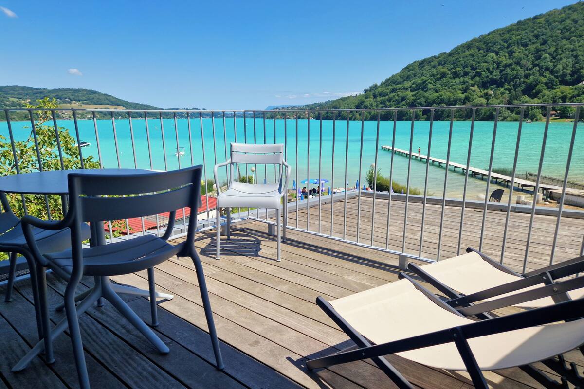 A wooden deck with two chairs and a table, overlooking a turquoise sea and a dock with boats.