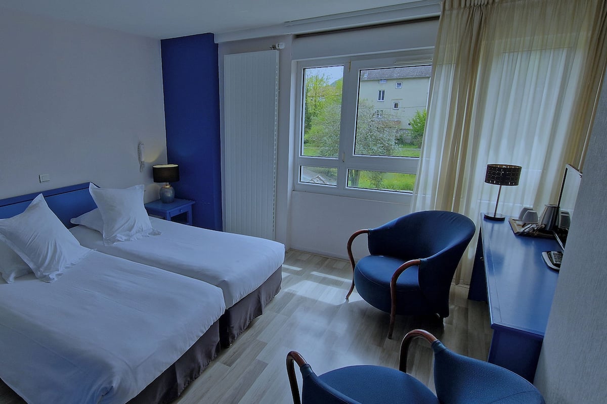 A hotel room with two beds, a desk, a chair, and a window with a view of greenery.