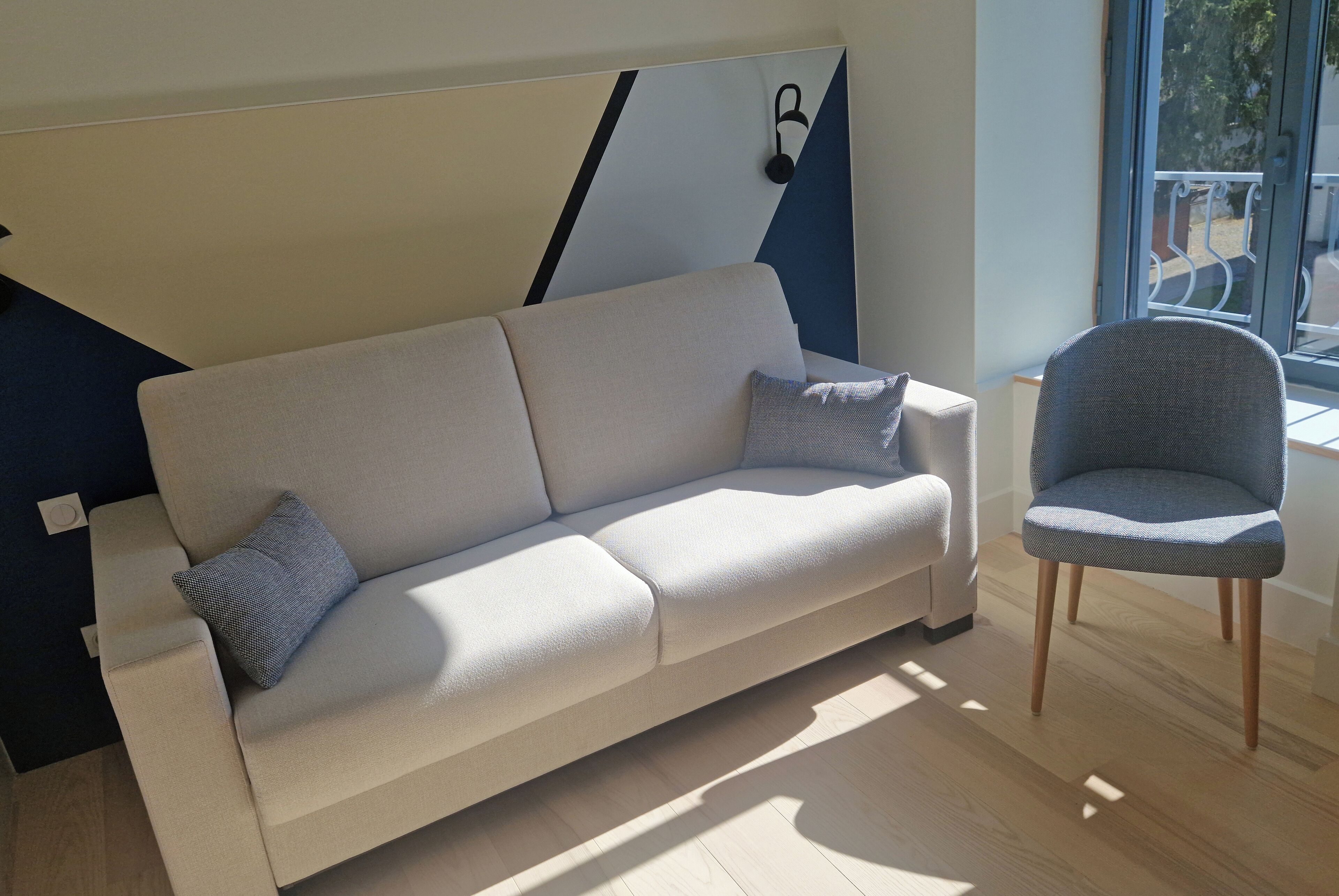 A modern living room with a sofa and a chair, a geometric wall design, and a view through a window.