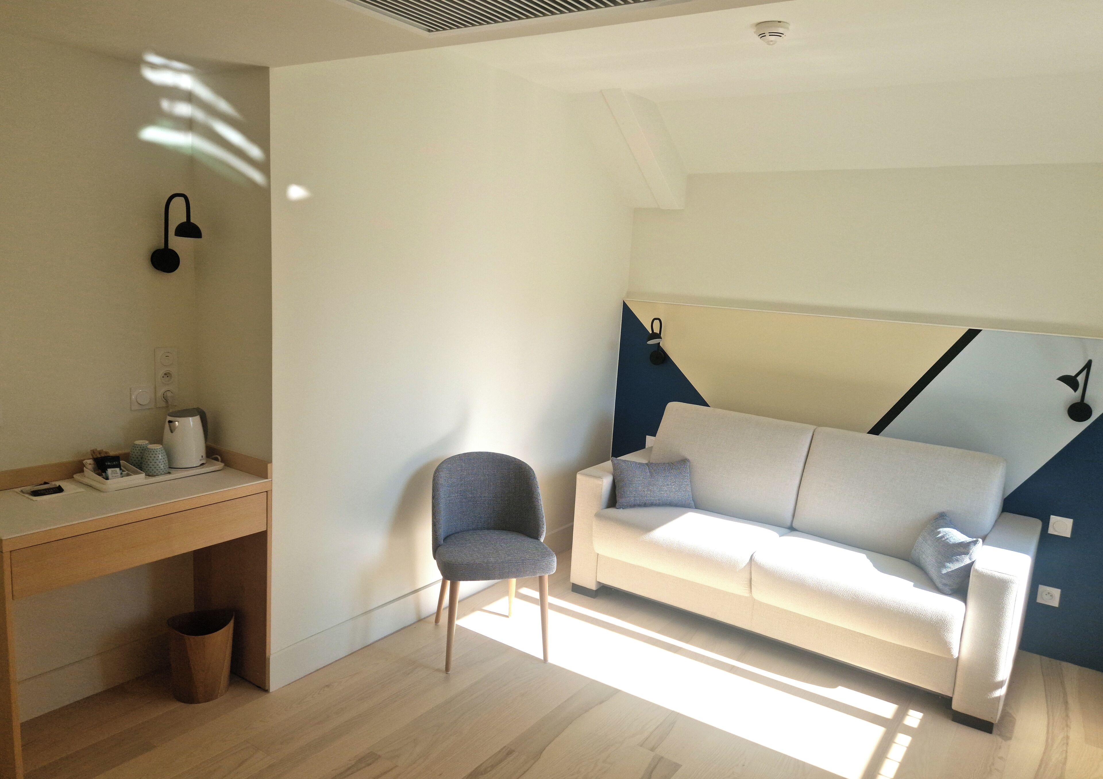 A modern, well-lit room with a wooden desk, a grey armchair, and a white sofa with blue accents.