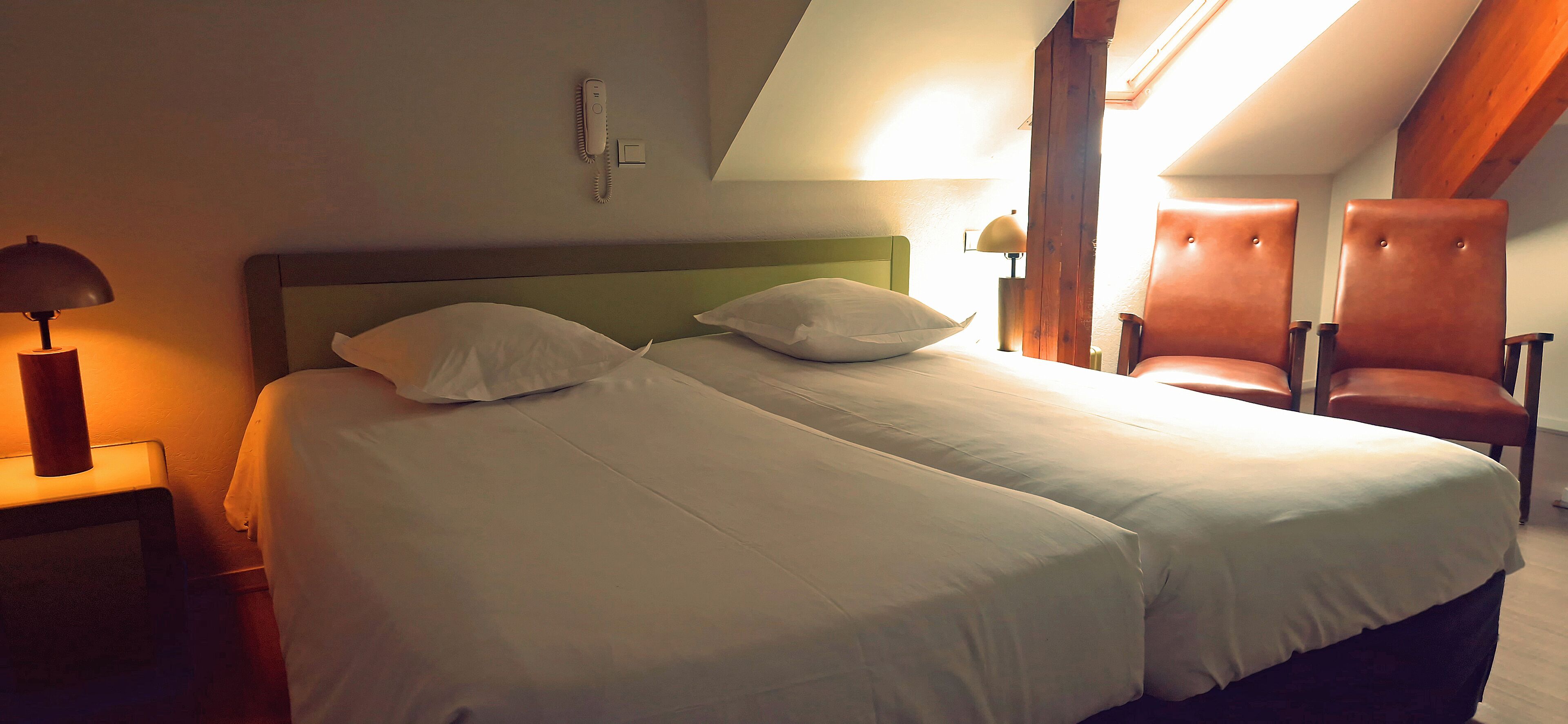 A double bed with white linens, a bedside lamp, a phone, and a chair in a room with wooden beams and a sloped ceiling.