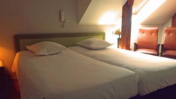 A double bed with white linens, a bedside lamp, a phone, and a chair in a room with wooden beams and a sloped ceiling.