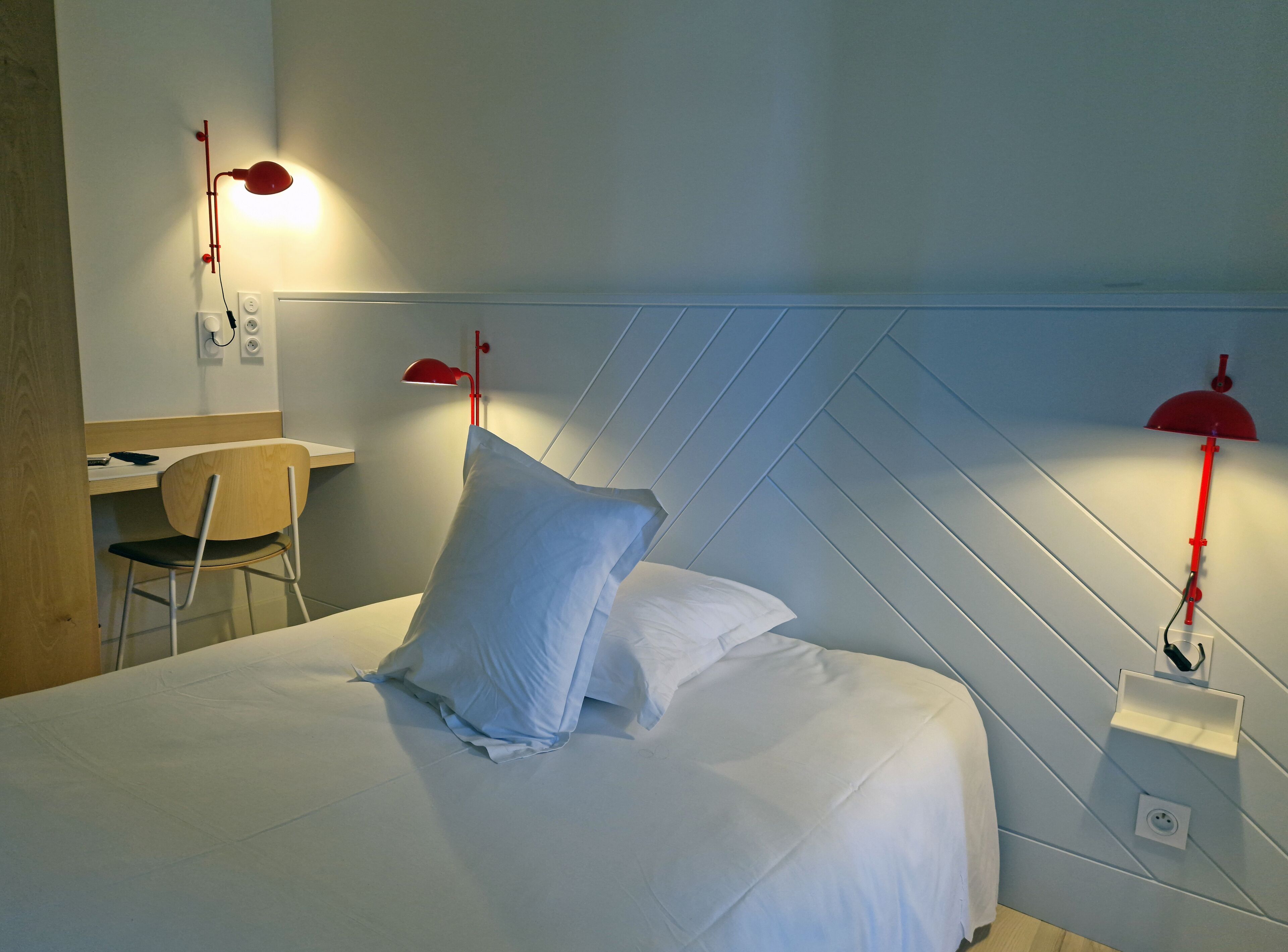 Una cama con sábanas blancas ordenadas, una silla de madera, un escritorio y dos lámparas de pared rojas.