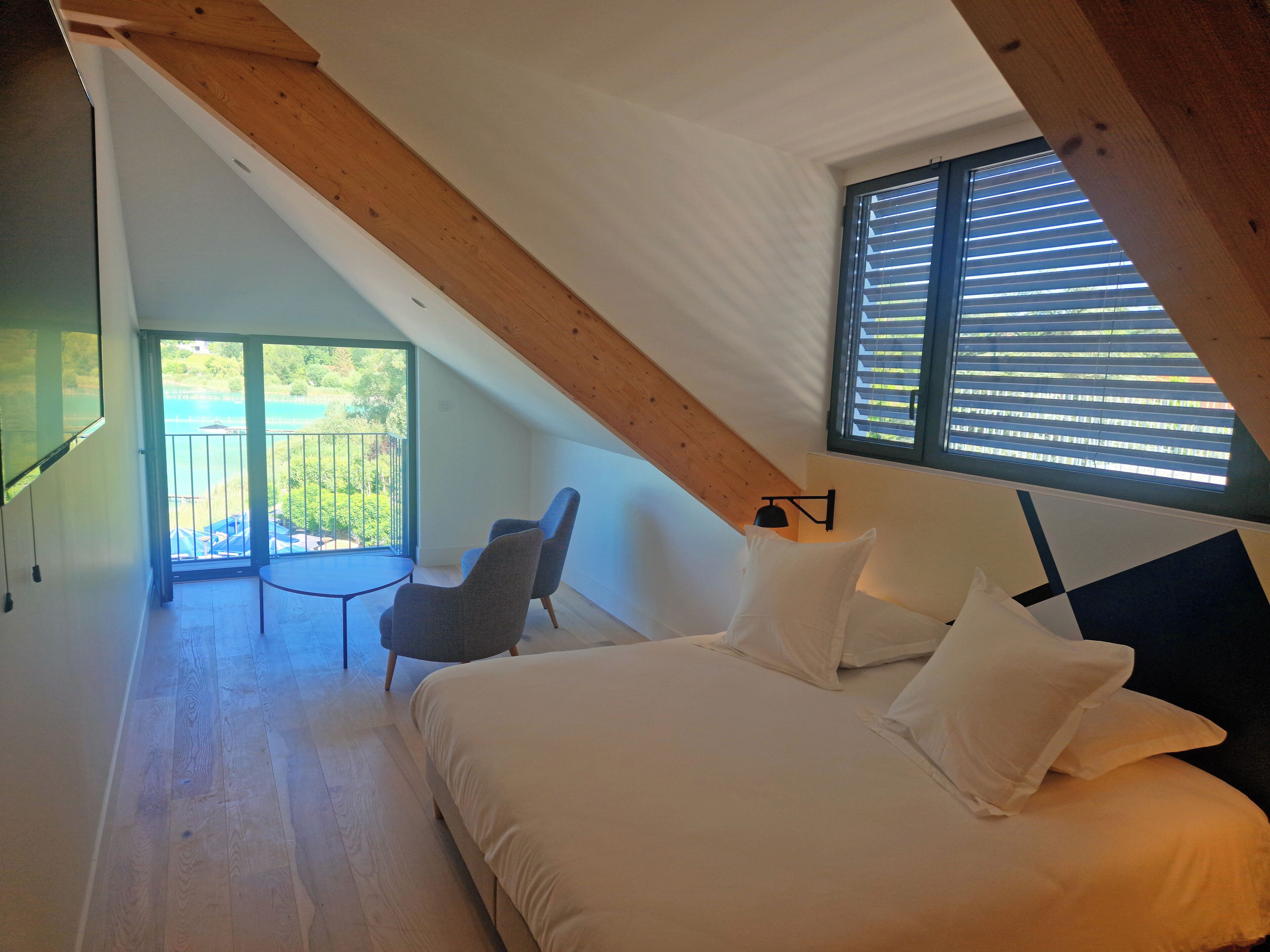 Een slaapkamer met een bed, twee stoelen, een tafeltje, een raam met zonwering en een balkon met uitzicht op groen.