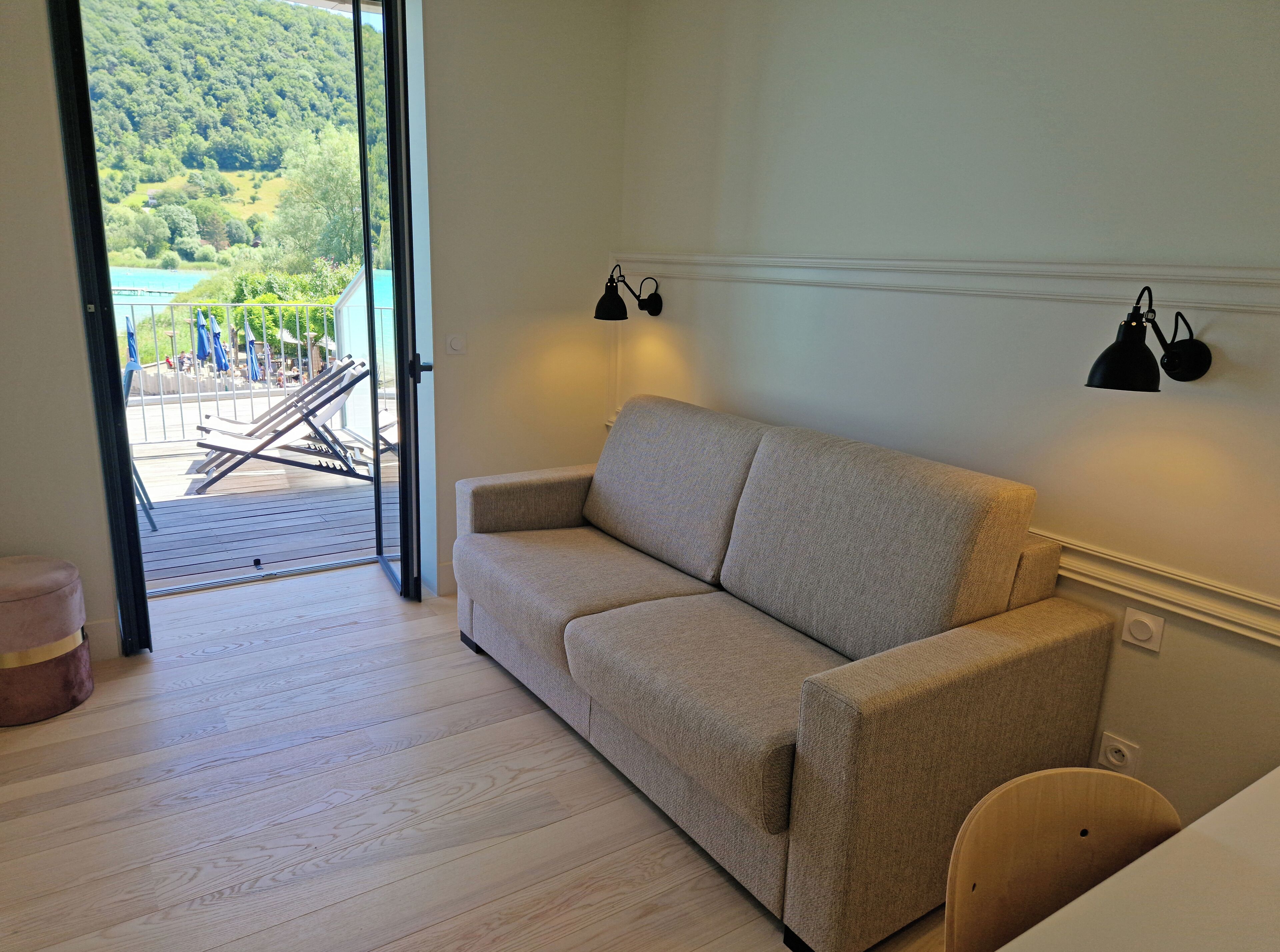 Una habitación con sofá color beige, piso de madera, puerta de vidrio que da a un área exterior con piscina y sillas de descanso, y dos lámparas fijadas a la pared.
