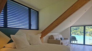 Un dormitorio con una cama grande, un sofá y una silla. Hay ventanas amplias con persianas, un balcón con barandilla y vistas a un lago.