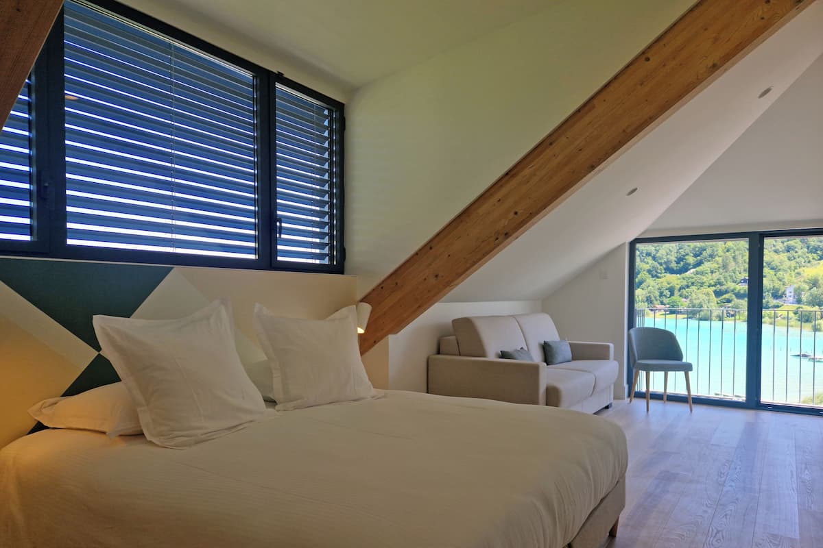 A bedroom with a large bed, a sofa, and a chair. There are large windows with blinds, a balcony with a railing, and a view of a lake.