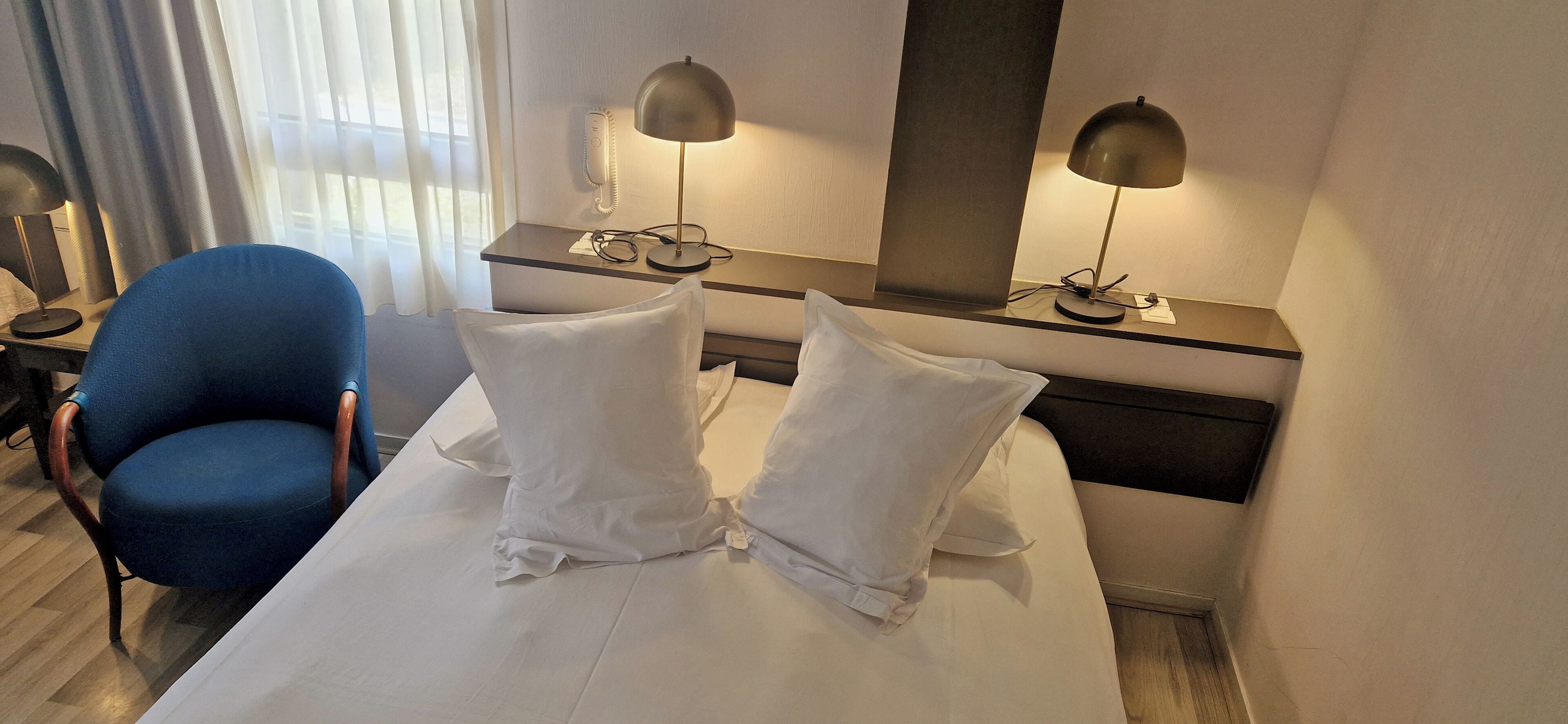 A hotel room with a bed, two bedside lamps, a desk with a telephone, a blue chair, and a window with curtains.