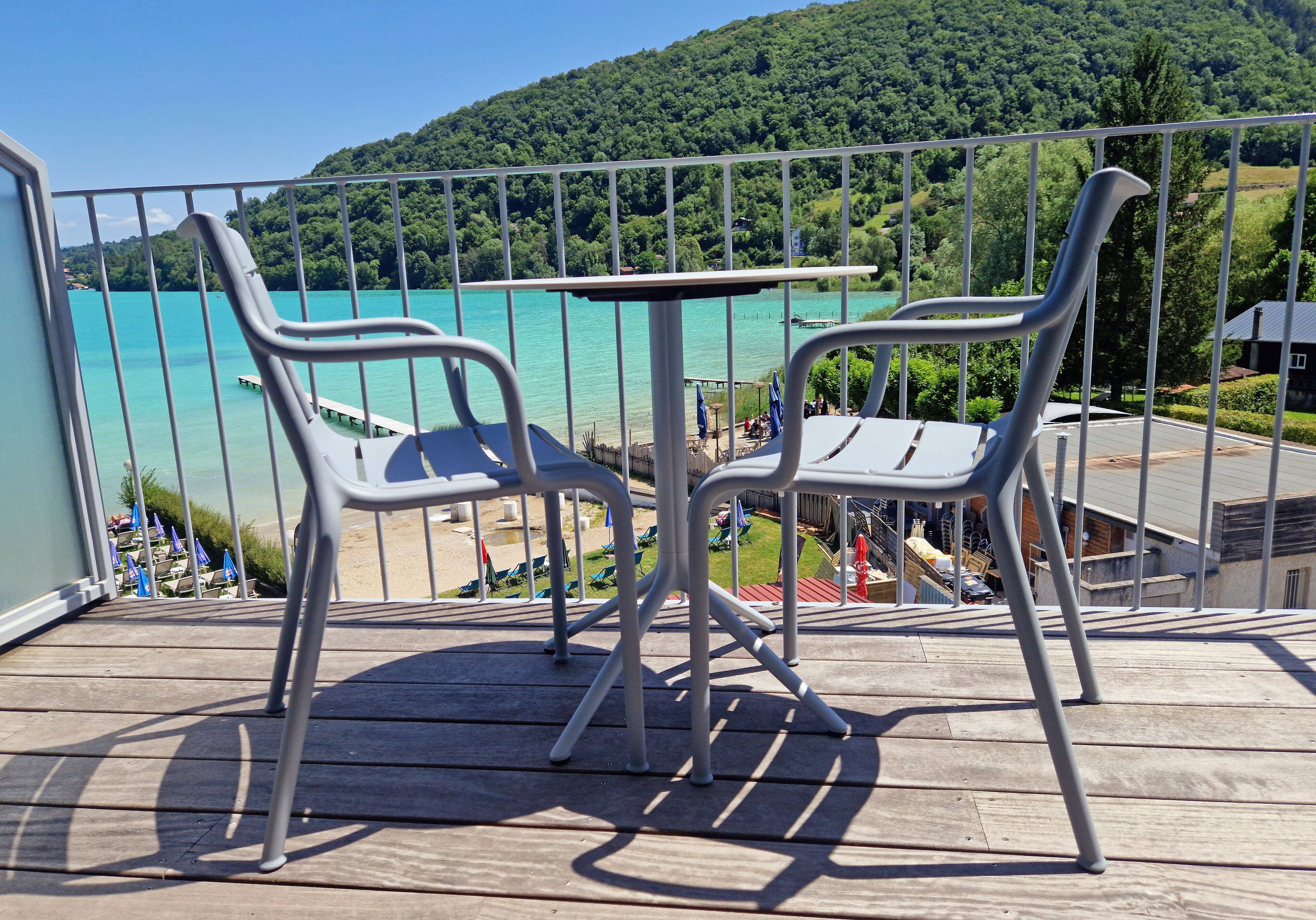 Un balcón con dos sillas de metal y una mesa, con vistas a un lago turquesa y una ladera boscosa.