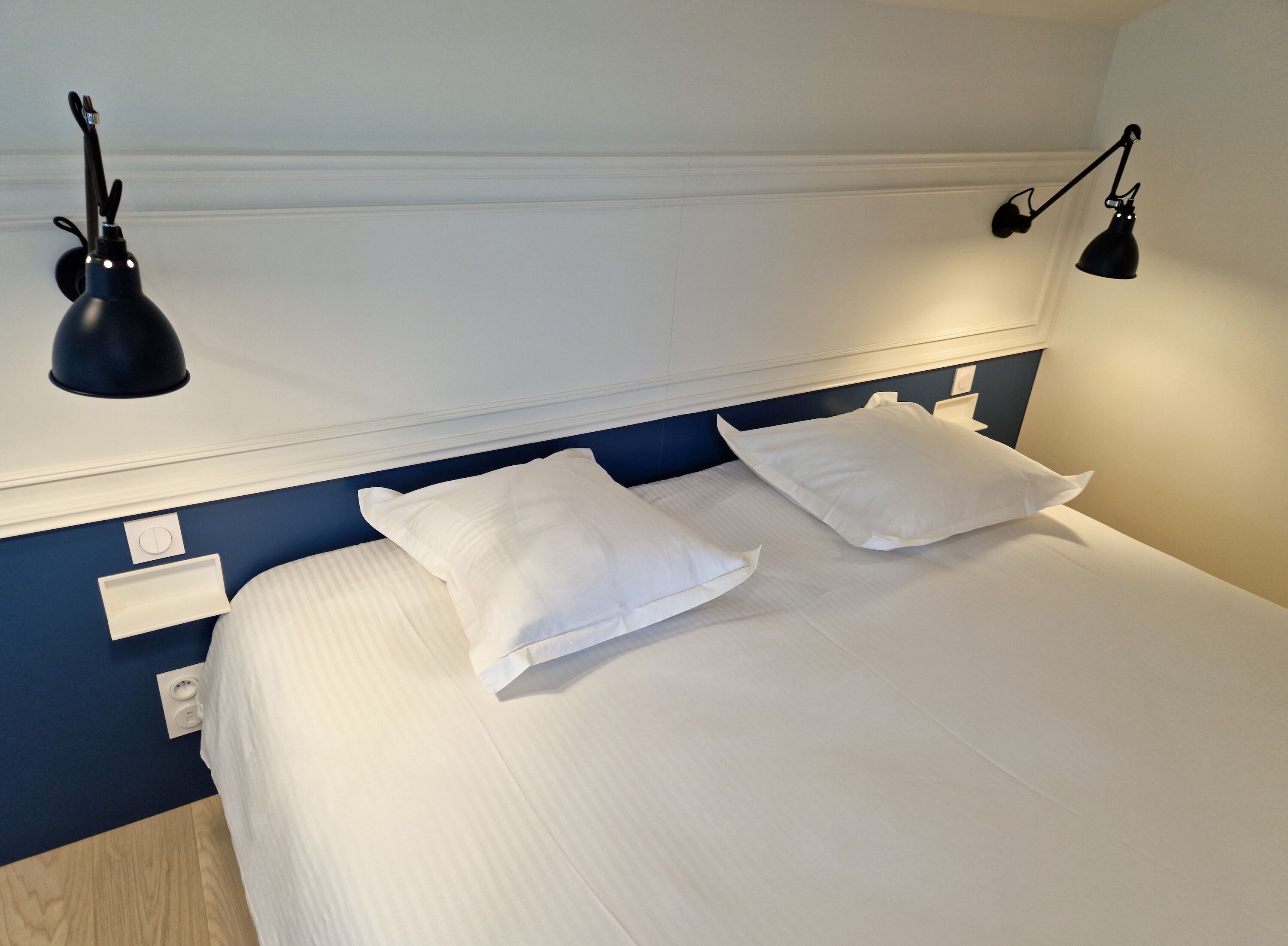 A hotel room with a neatly made bed, two bedside lamps, and a wall-mounted light fixture.