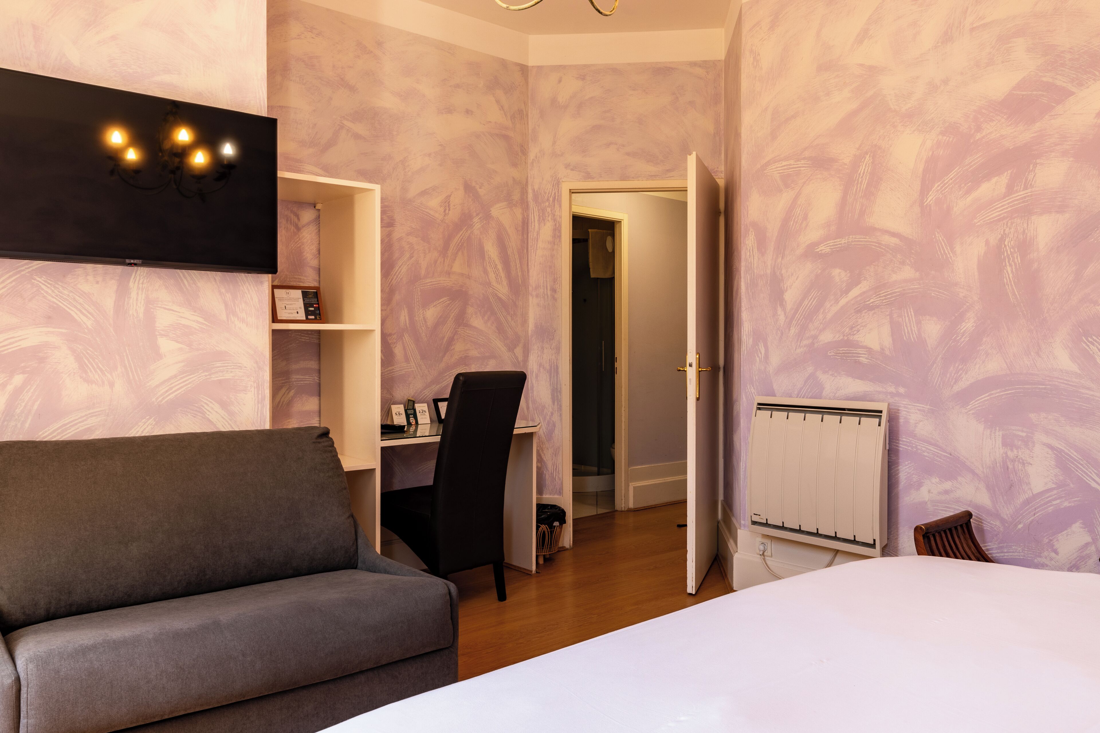A hotel room with a grey sofa, a desk with a chair, a TV mounted on the wall, and a radiator.