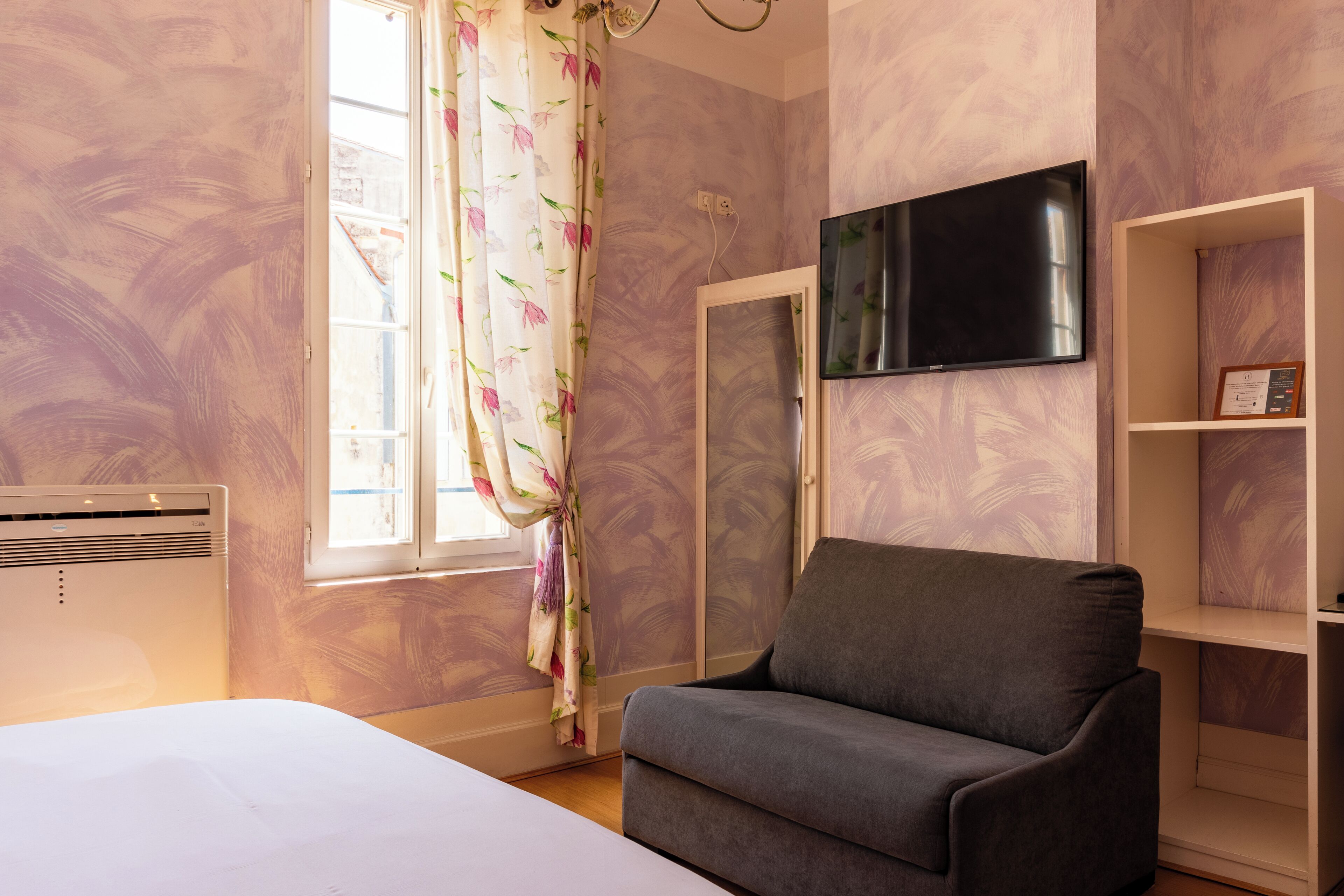 A room with a bed, a grey sofa, a TV mounted on the wall, and a window with floral curtains.