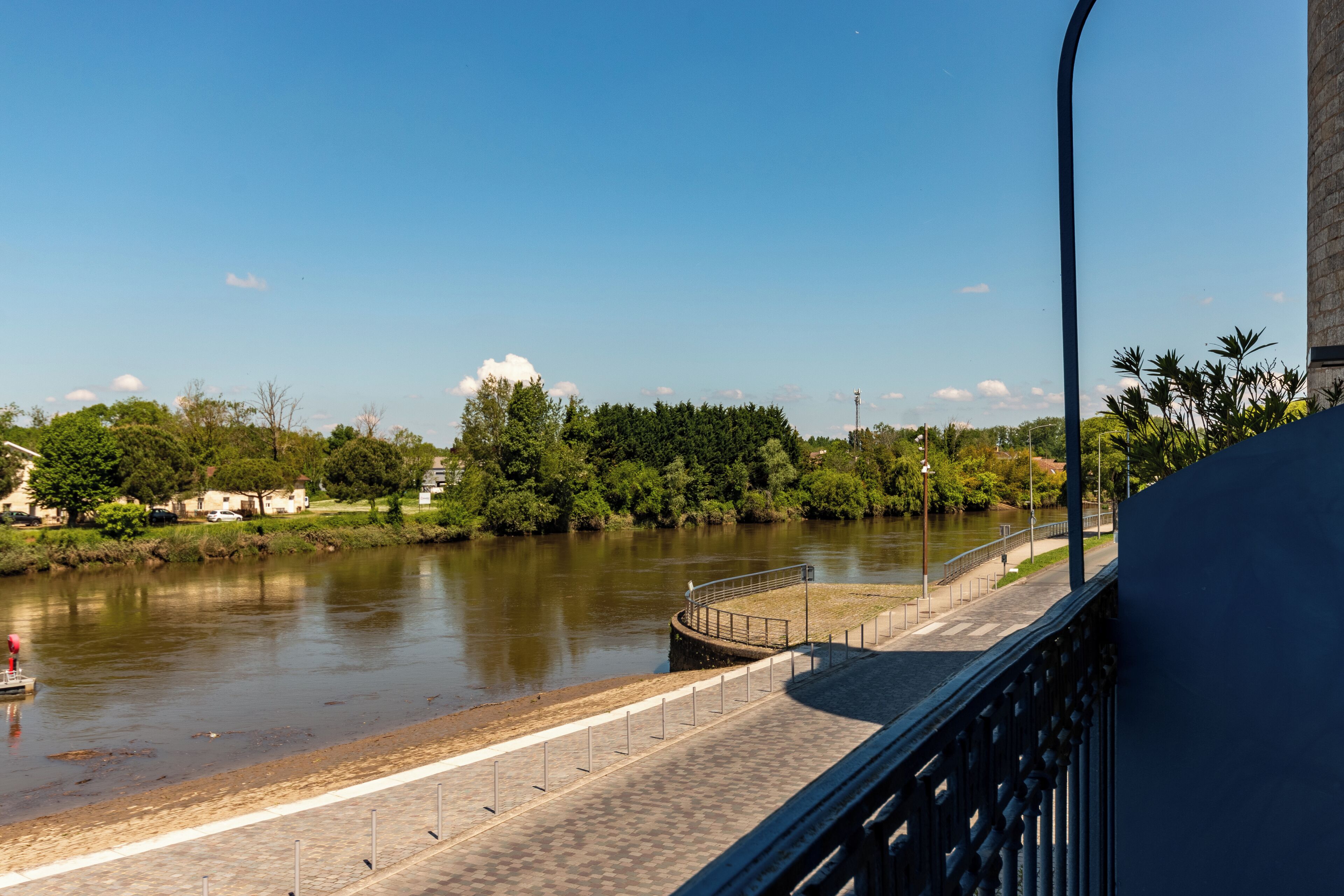 A riverside walkway with a paved path, a railing, and a view of a river and greenery.