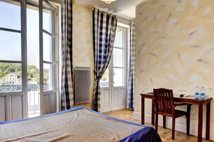 Una habitación con cama, un escritorio de madera y una silla. Cuenta con ventanas grandes con cortinas, un radiador y vistas a edificios.