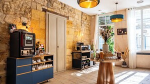 A kitchen with a stone wall, a coffee machine, a coffee cart, and a round wooden table with a vase of dried plants. - Hôtel de la tour à LIBOURNE (Libourne)