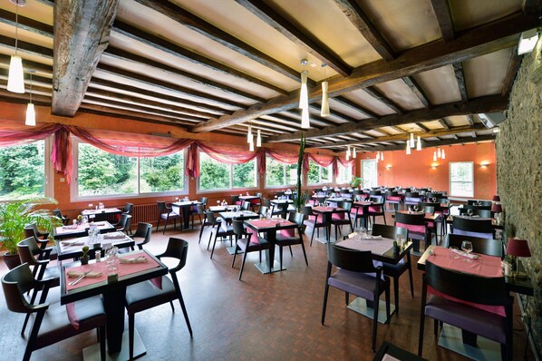 A spacious restaurant with multiple tables and chairs, large windows with red drapes, and hanging pendant lights. - Contact Hôtel le Moulin Neuf (Chantonnay)