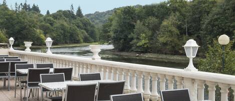 A terrace with tables and chairs, overlooking a river and forested hills.