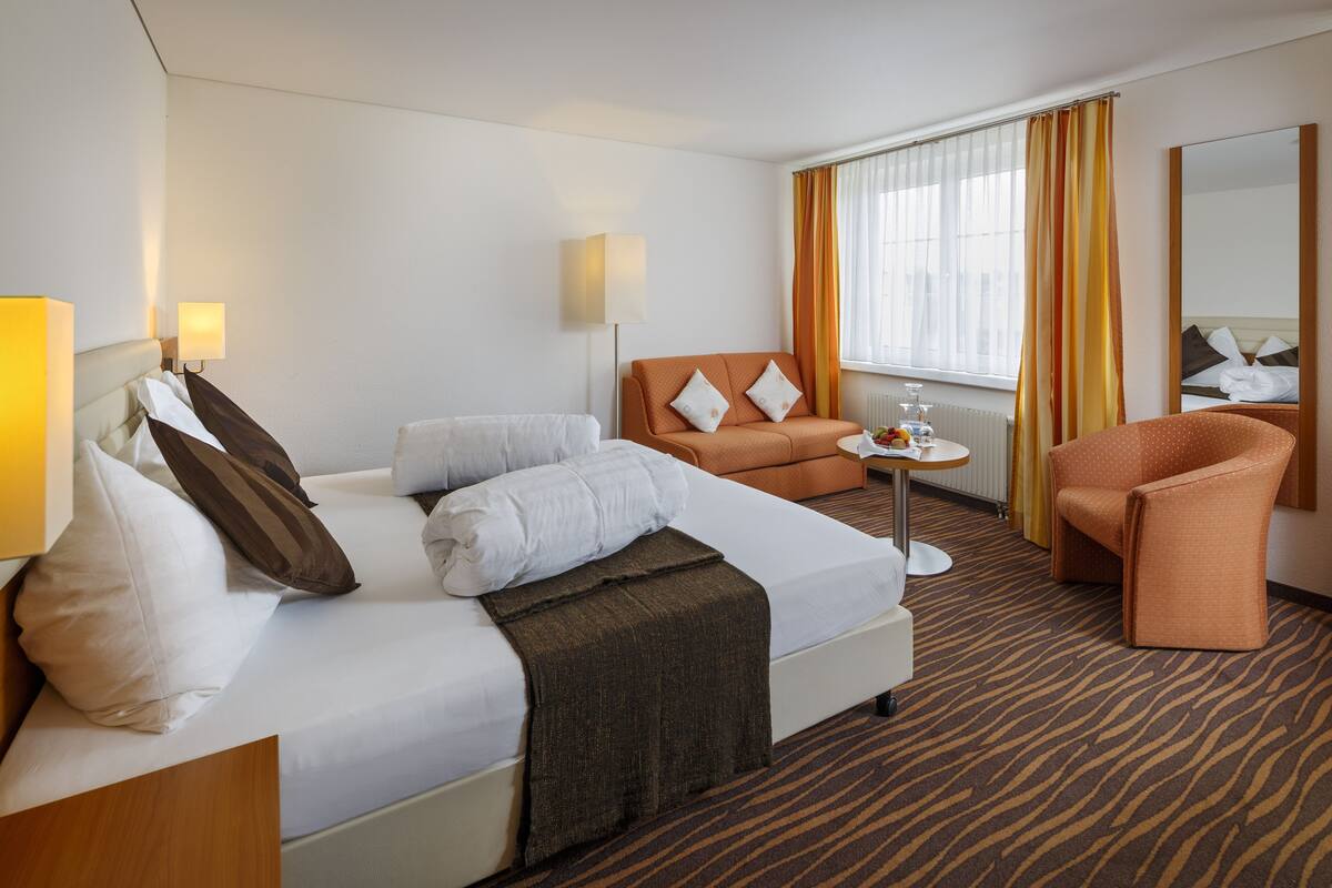 Une chambre d’hôtel avec un lit, un canapé, une chaise, une petite table avec un vase et une fenêtre avec des rideaux.