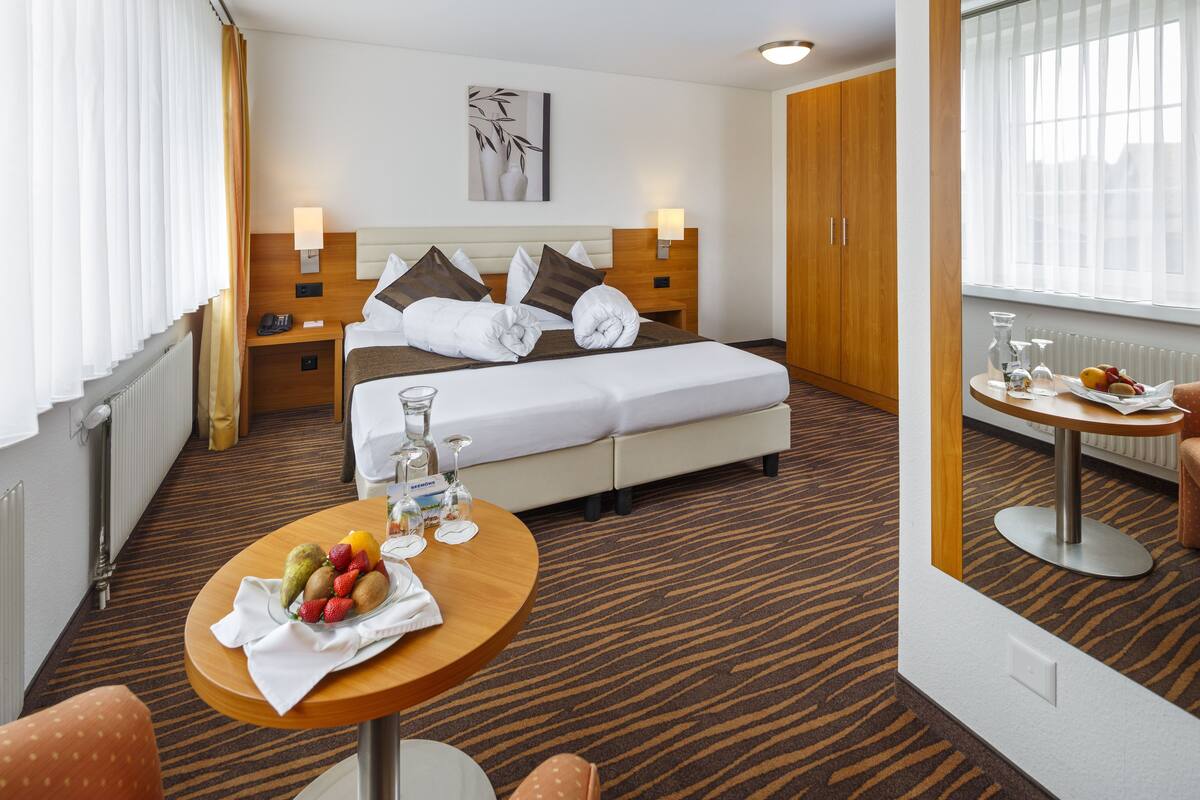 Une chambre d’hôtel avec un lit, une table de chevet sur laquelle se trouve un panier de fruits, une table ronde également surmontée d’un panier de fruits, un miroir et une fenêtre avec des rideaux.