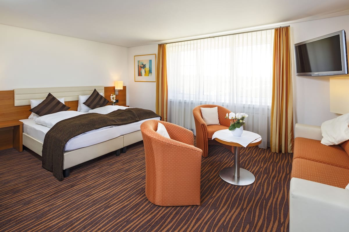 Une chambre d’hôtel comprenant un lit, deux fauteuils, une petite table, un téléviseur et une fenêtre avec des rideaux.