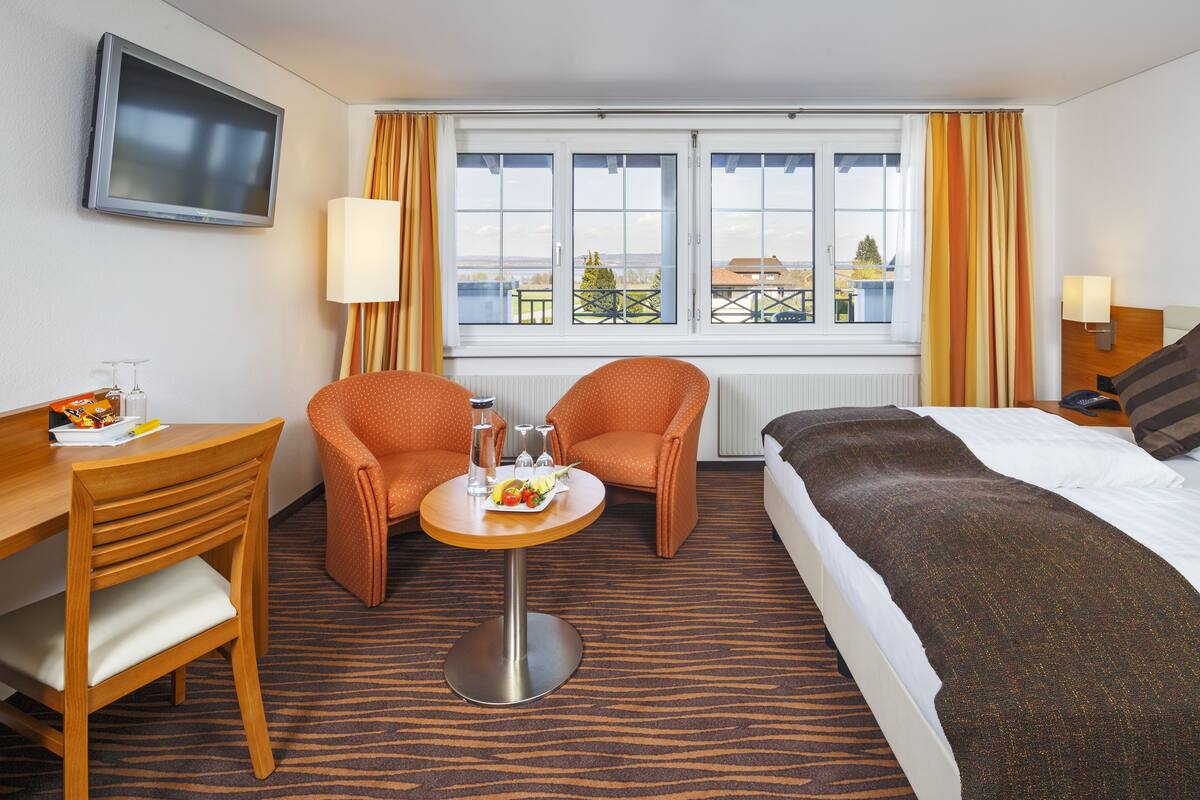 Une chambre d’hôtel comprenant un lit, deux fauteuils, une petite table avec une assiette de fruits, un téléviseur fixé au mur et une fenêtre avec des rideaux.