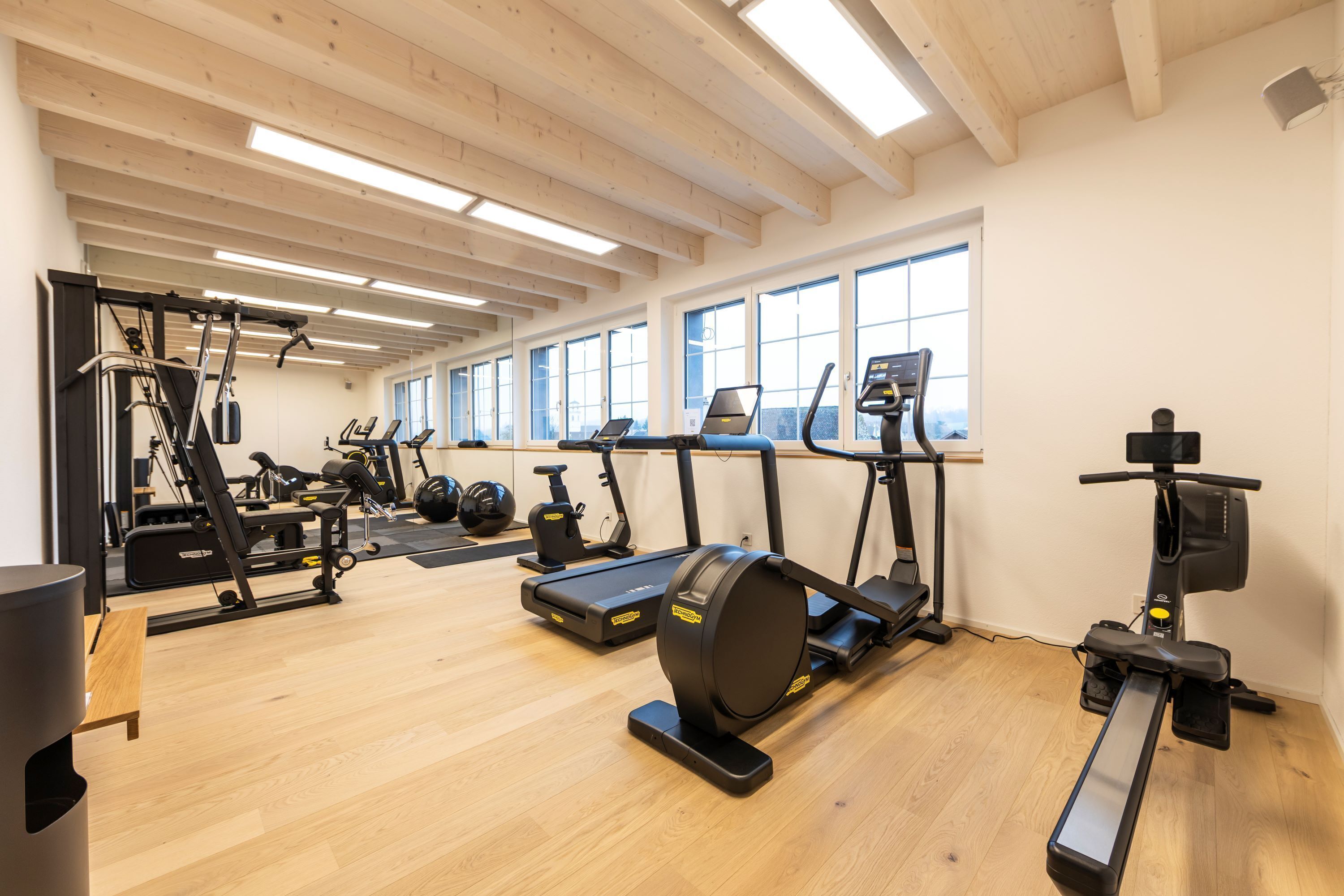 Un gimnasio con diversos equipos de ejercicio, amplias ventanas y suelos de madera.