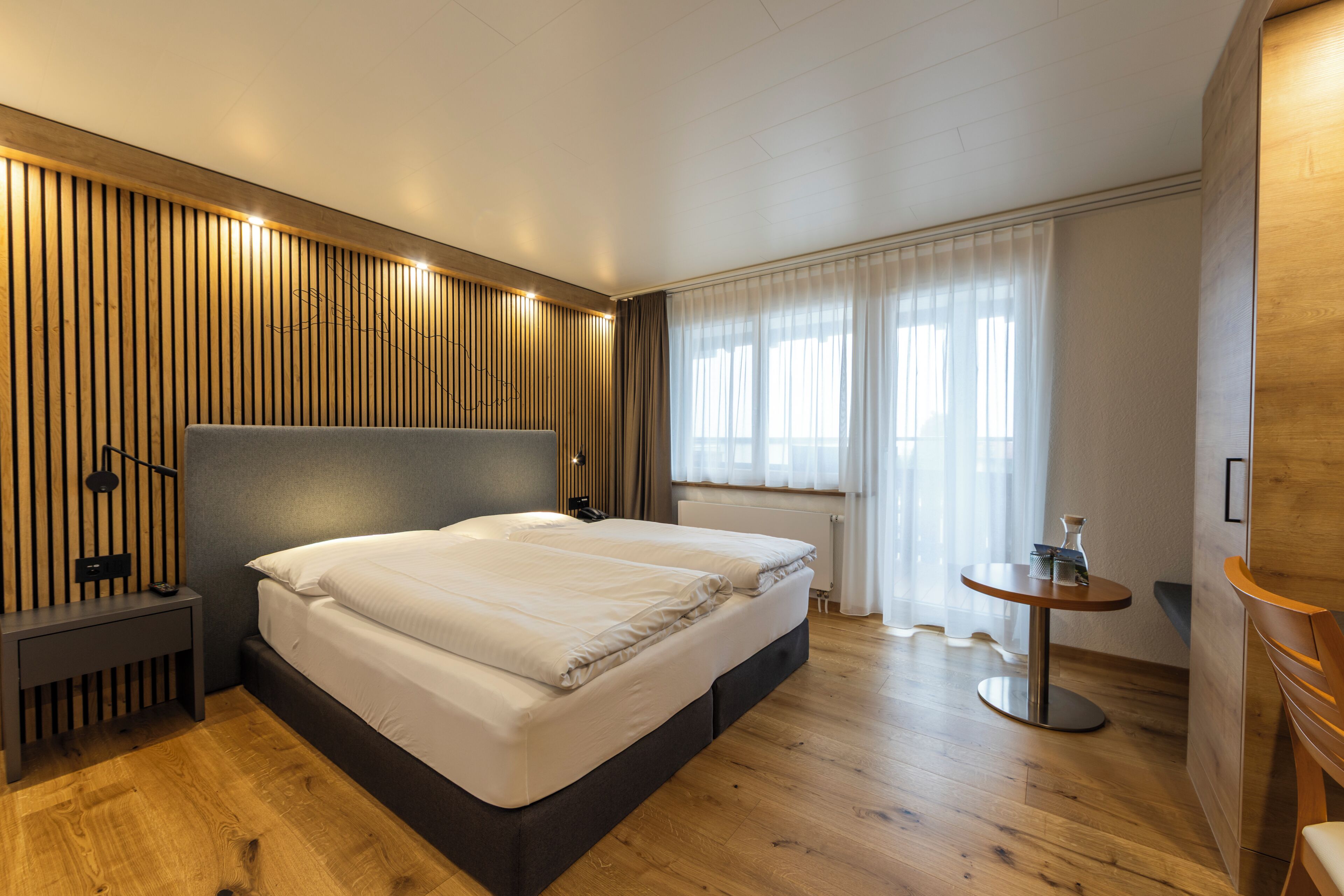 Moderni hotellihuone, jossa on suuri sänky, yöpöydät, pieni pyöreä pöytä ja ikkuna, jossa on verhot.
