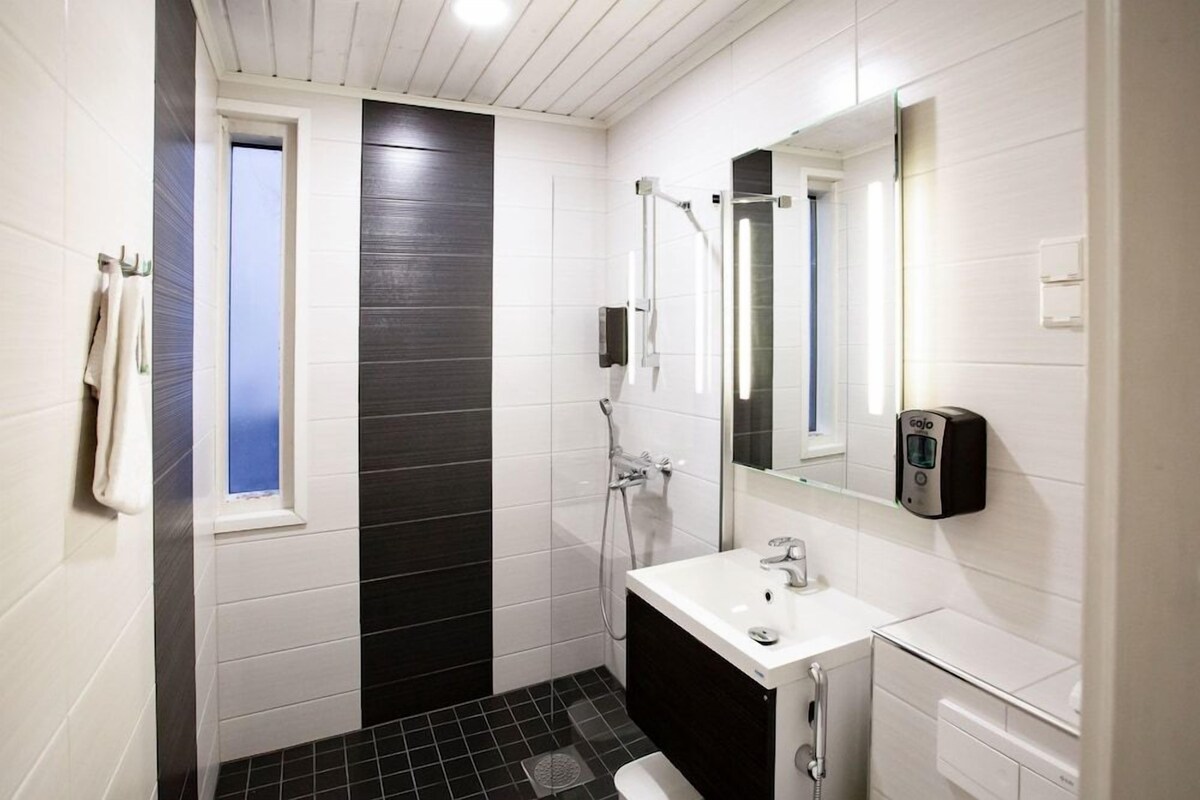Baño con lavamanos blanco, espejo, cabezal de ducha de mano y toallero.