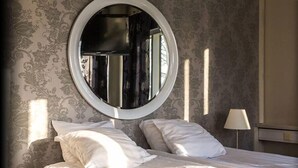 Un dormitorio con un espejo redondo, dos almohadas blancas, una lámpara de noche y papel tapiz estampado.