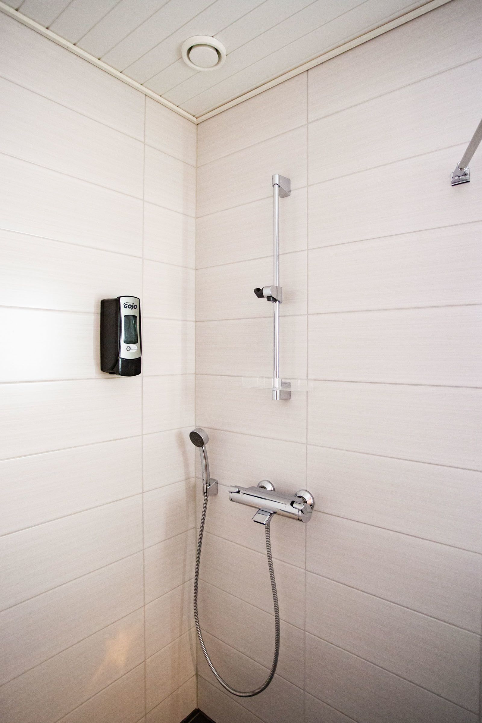 A bathroom with white tiled walls, a handheld showerhead, a wall-mounted faucet, and a soap dispenser.