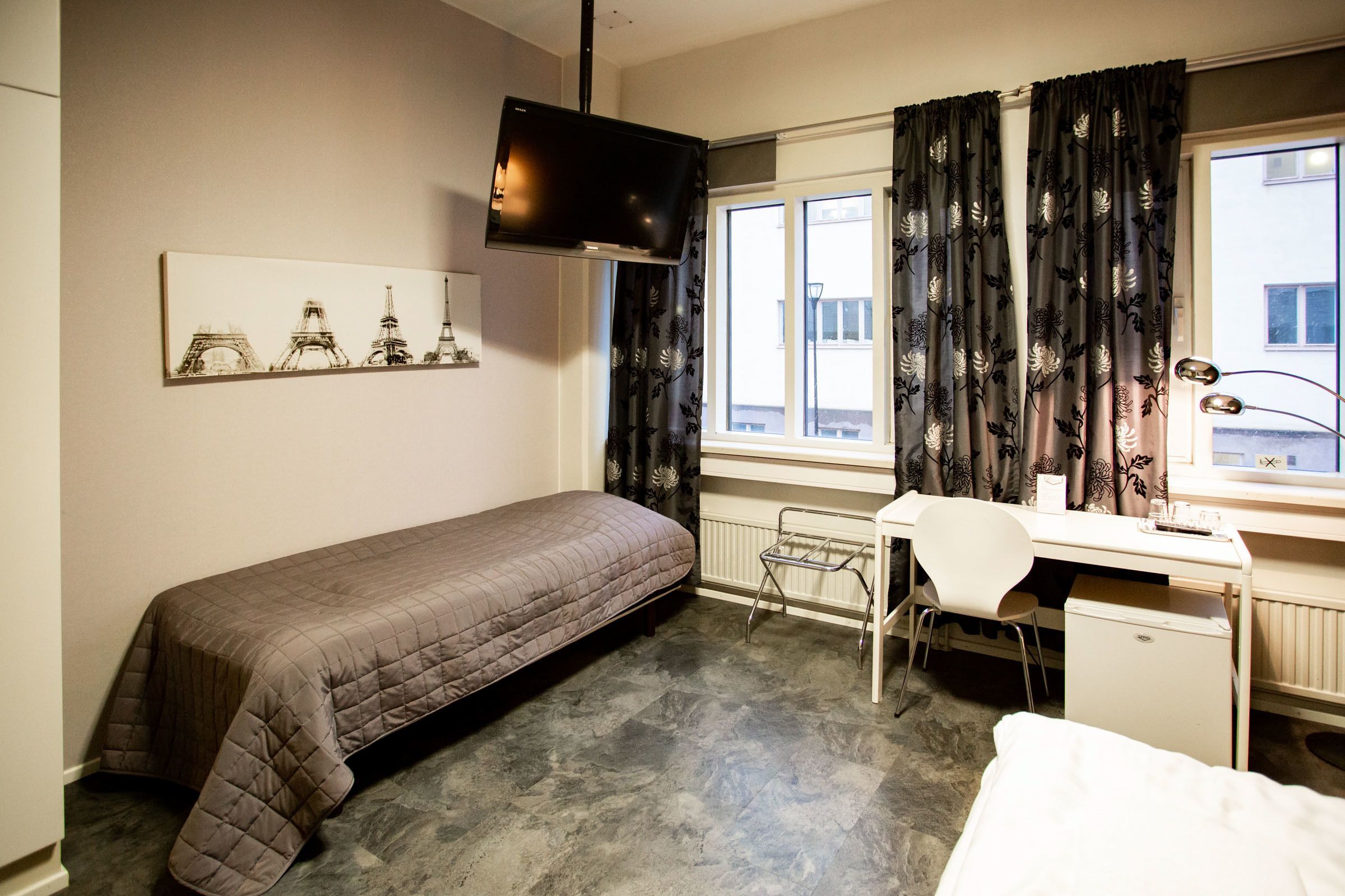 A bedroom with a bed, a desk, a chair, a radiator, a TV, and a window with curtains.