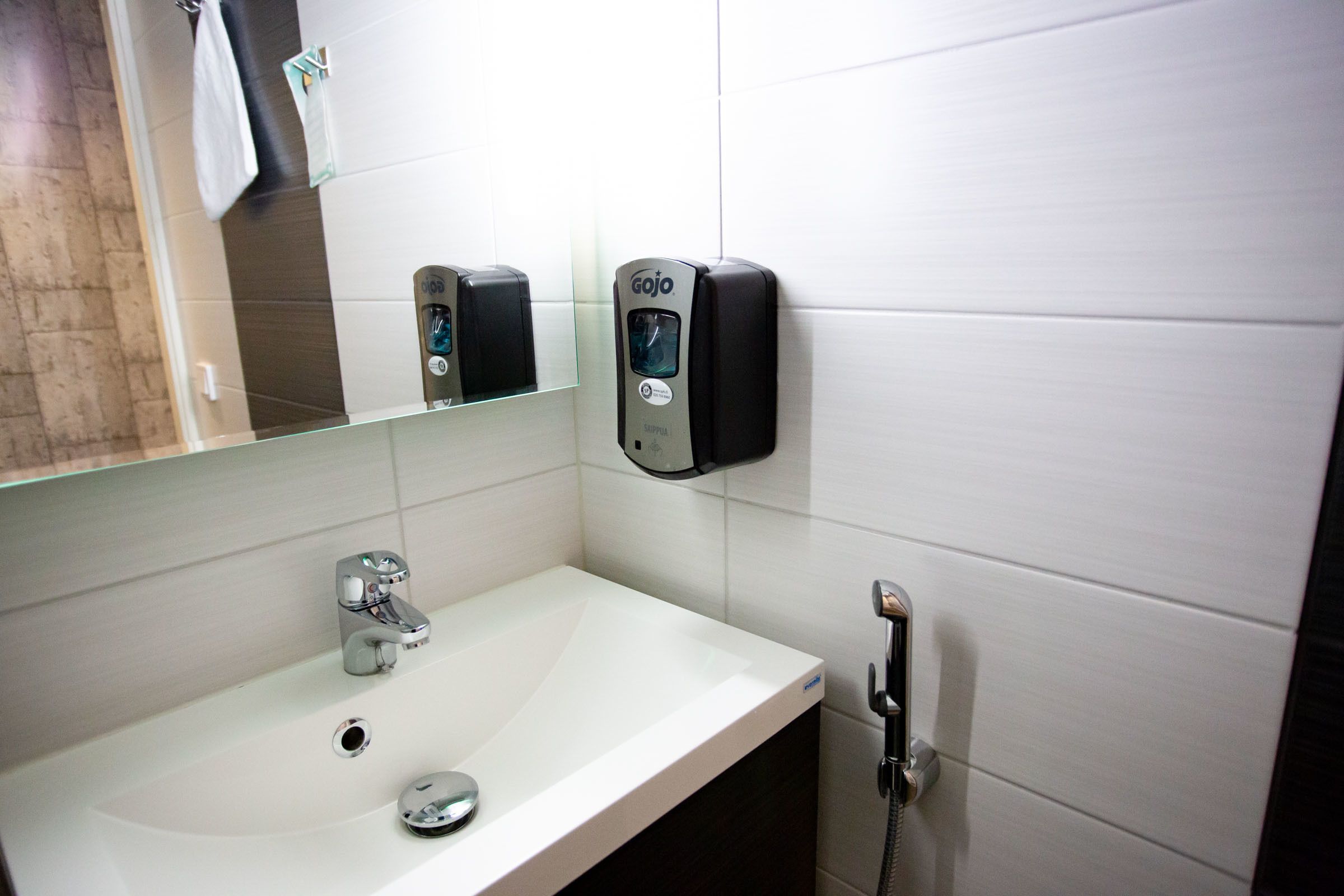Un baño con lavamanos blanco, espejo, dispensador de jabón y cabezal de ducha de mano.