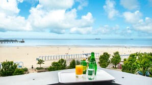 Mesa con bandeja que contiene un vaso de jugo de naranja y dos botellas, con vista a una playa y un muelle, y un barco a lo lejos.