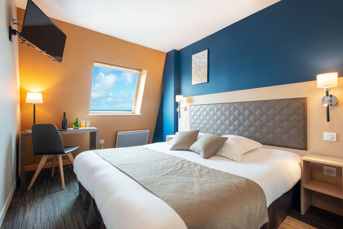 Une chambre d’hôtel équipée d’un lit, d’un bureau, d’une chaise et d’une fenêtre donnant sur le ciel.