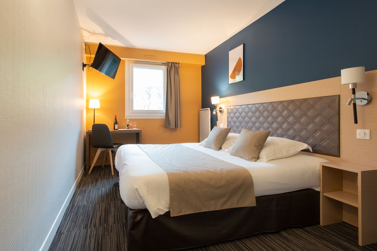 Une chambre d’hôtel avec un grand lit, un bureau avec une chaise, un téléviseur fixé au mur et une fenêtre avec des rideaux.