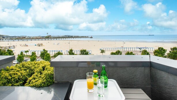 Un balcón con una bandeja que contiene un vaso de zumo de naranja, un vaso de agua y una botella verde, con vistas a una playa con sombrillas y un muelle.