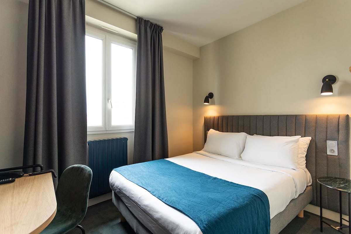 A hotel room with a bed, a desk, a chair, and a window with curtains.