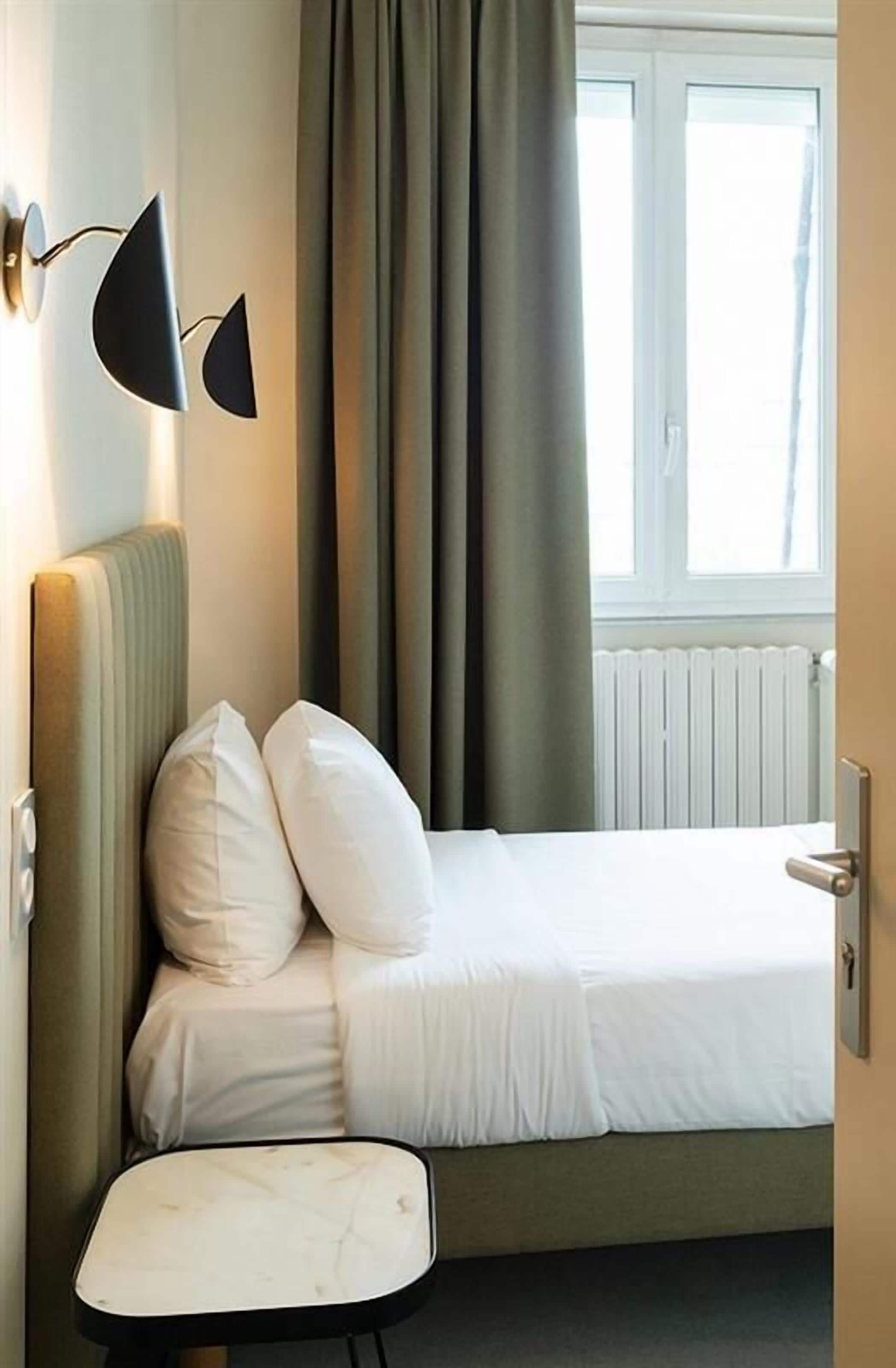 A small, neatly made bed with white linens and two pillows, a small table with a white top, and a wall-mounted lamp.