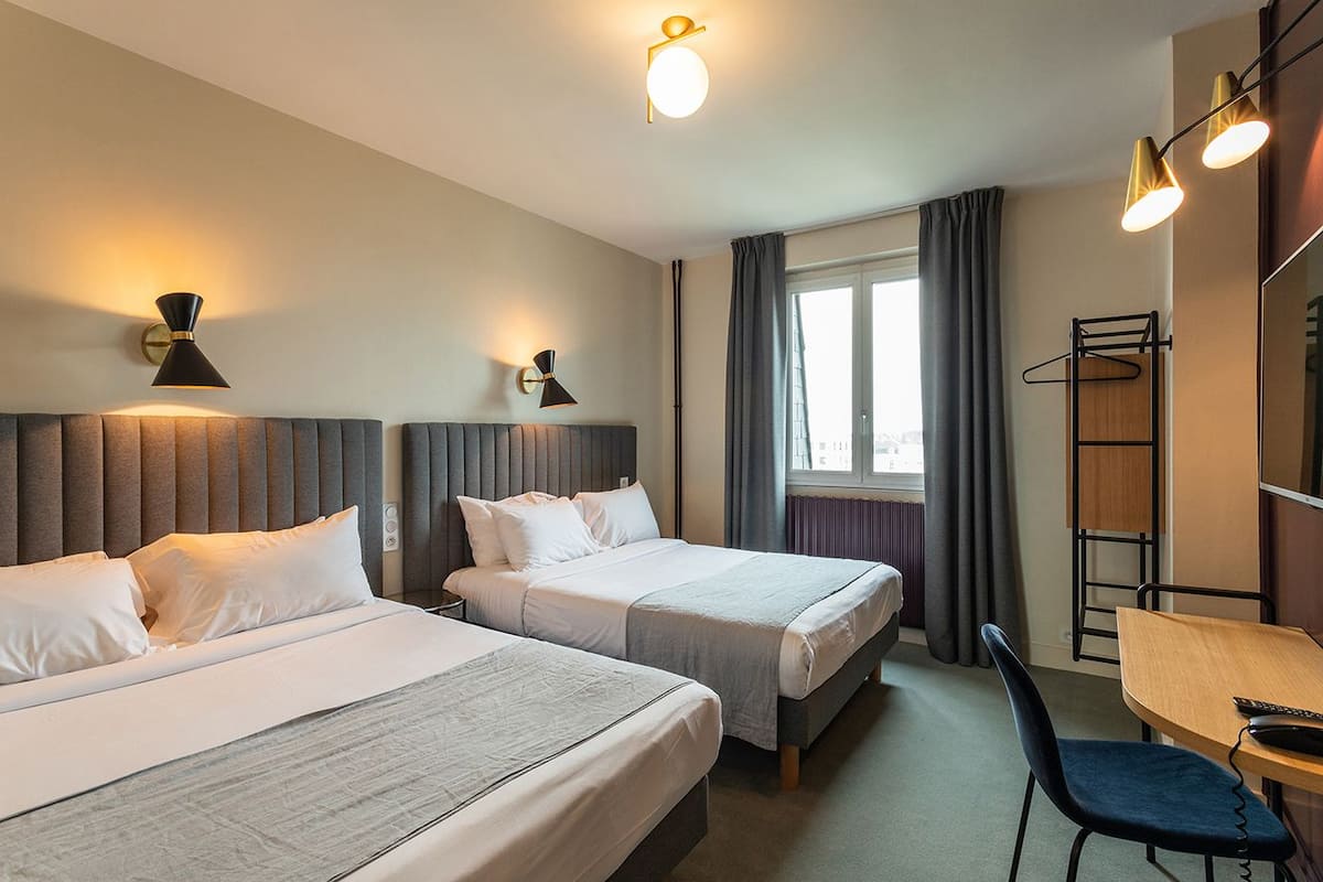 A hotel room with two beds, a desk, and a chair. There are wall-mounted lamps and a window with curtains.