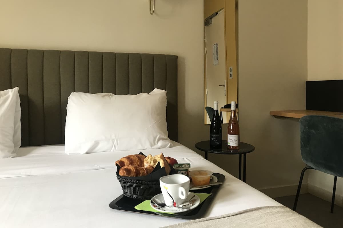 A hotel room with a bed, a bedside table with a bottle of wine, a basket of croissants, a cup of coffee, and a chair.