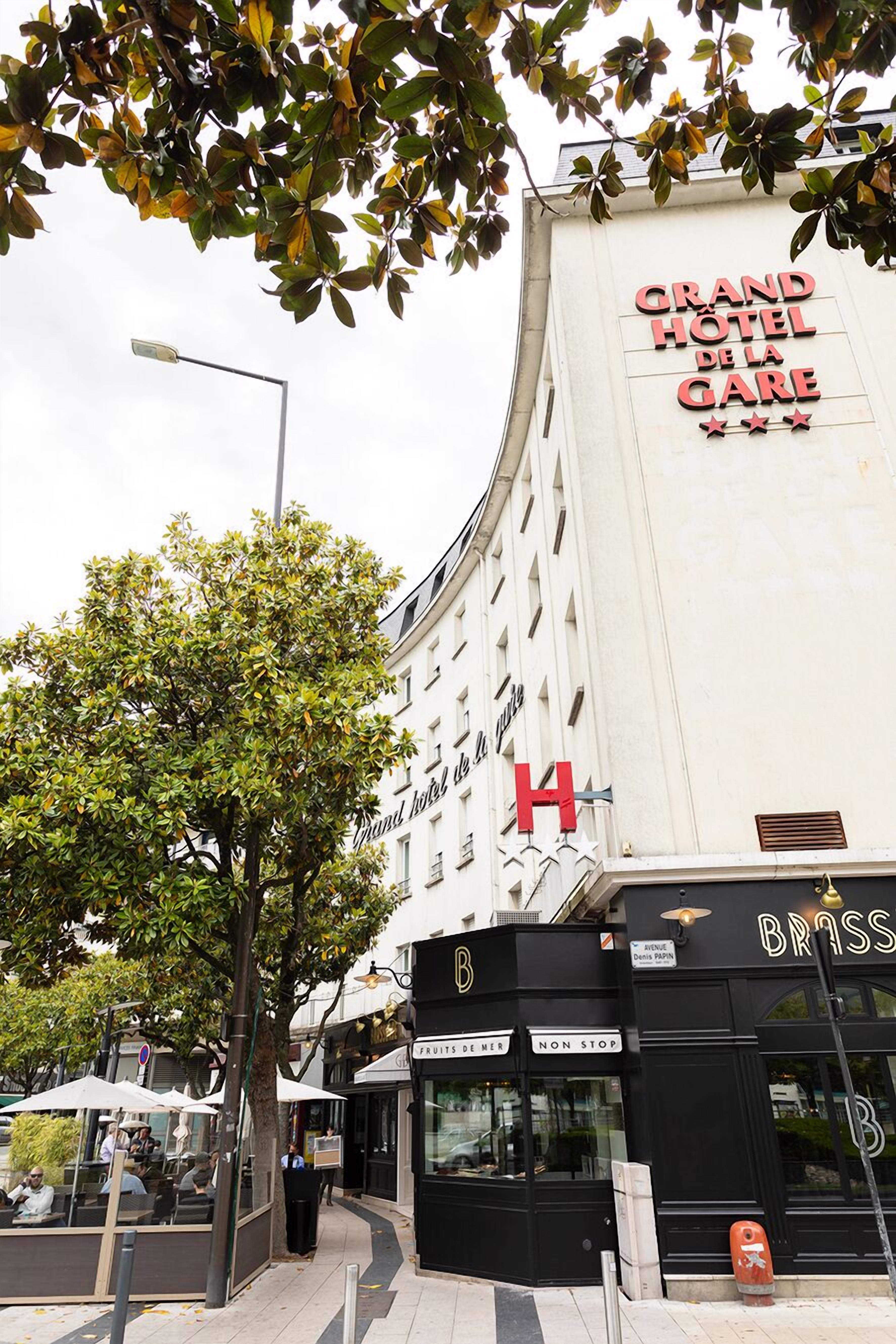 A hotel with a prominent 'Grand Hotel de la Gare' sign, a 'Brass' storefront, and outdoor seating with umbrellas.