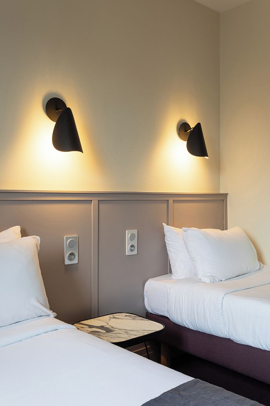 A hotel room with two beds, a small table, and wall-mounted lamps.