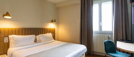 Habitación de hotel con cama, lámparas de noche, una silla, una mesa y un ventanal con cortinas.