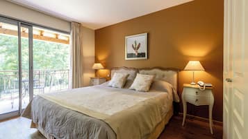 Une chambre avec un grand lit, deux tables de chevet avec des lampes, un tableau encadré au mur et un balcon avec du mobilier d’extérieur.